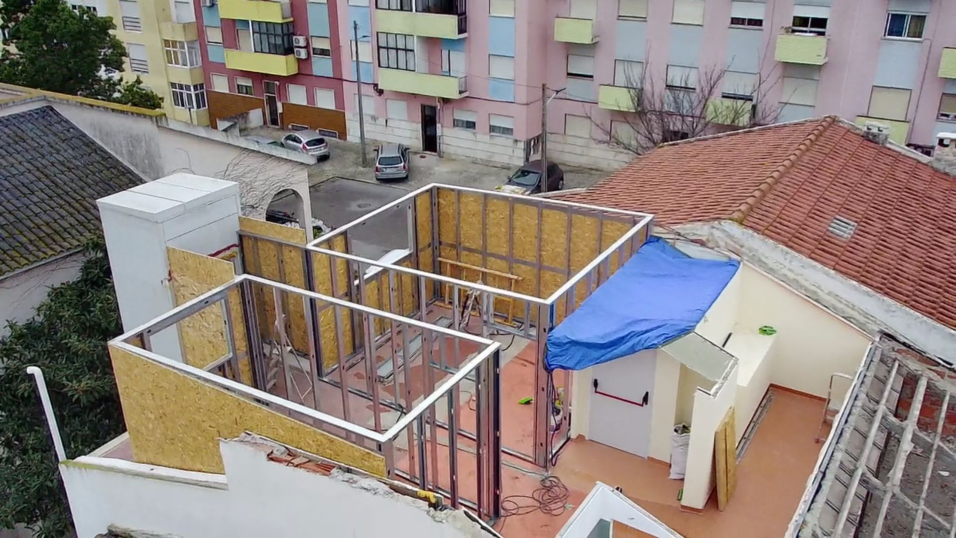 Vista aérea de um canteiro de obras com paredes estruturadas em um telhado, coberto por uma lona azul.