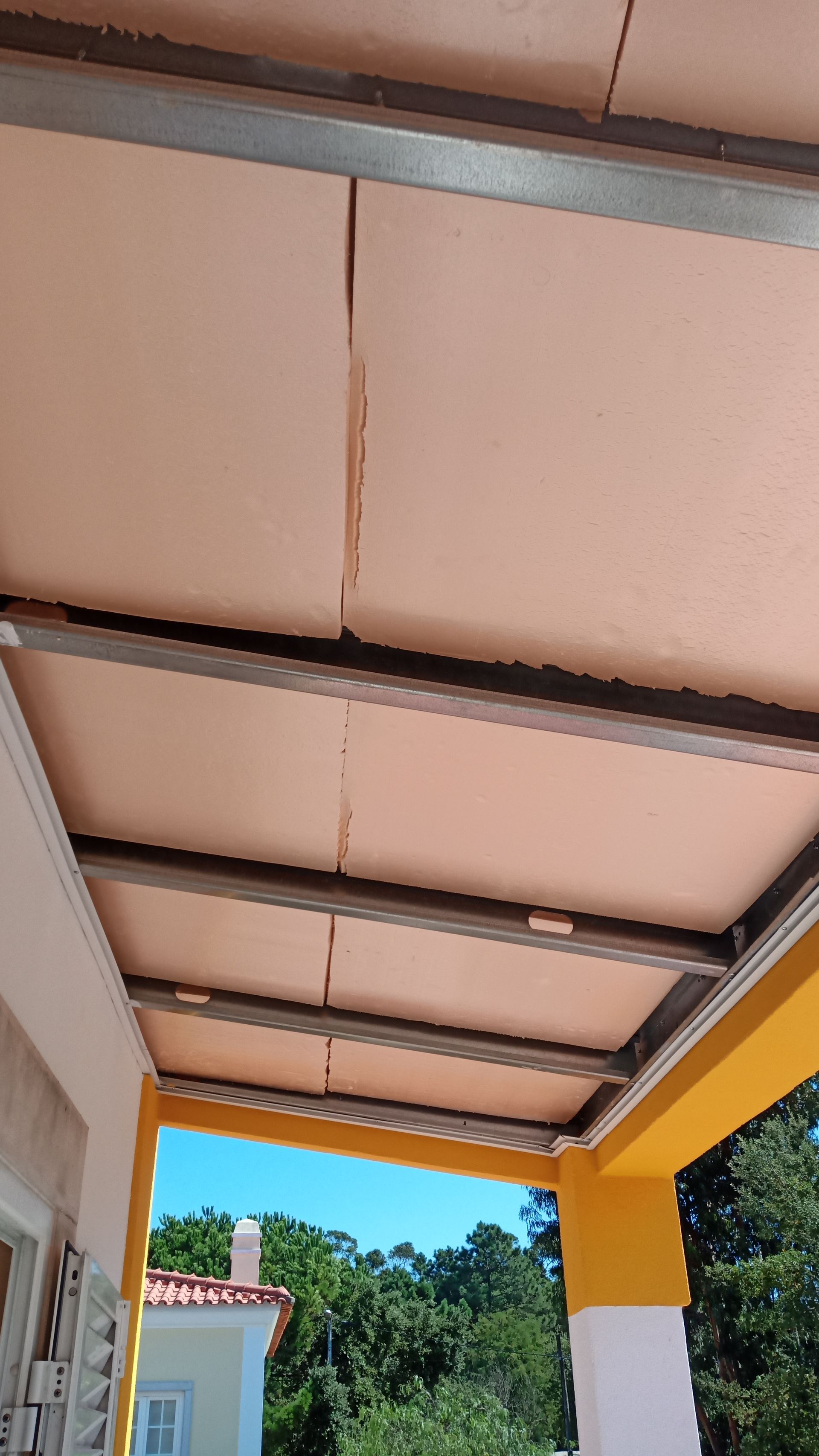 Painéis de toldo cor de bege fixados a uma estrutura metálica no telhado, sobre um espaço externo com colunas amarelas.