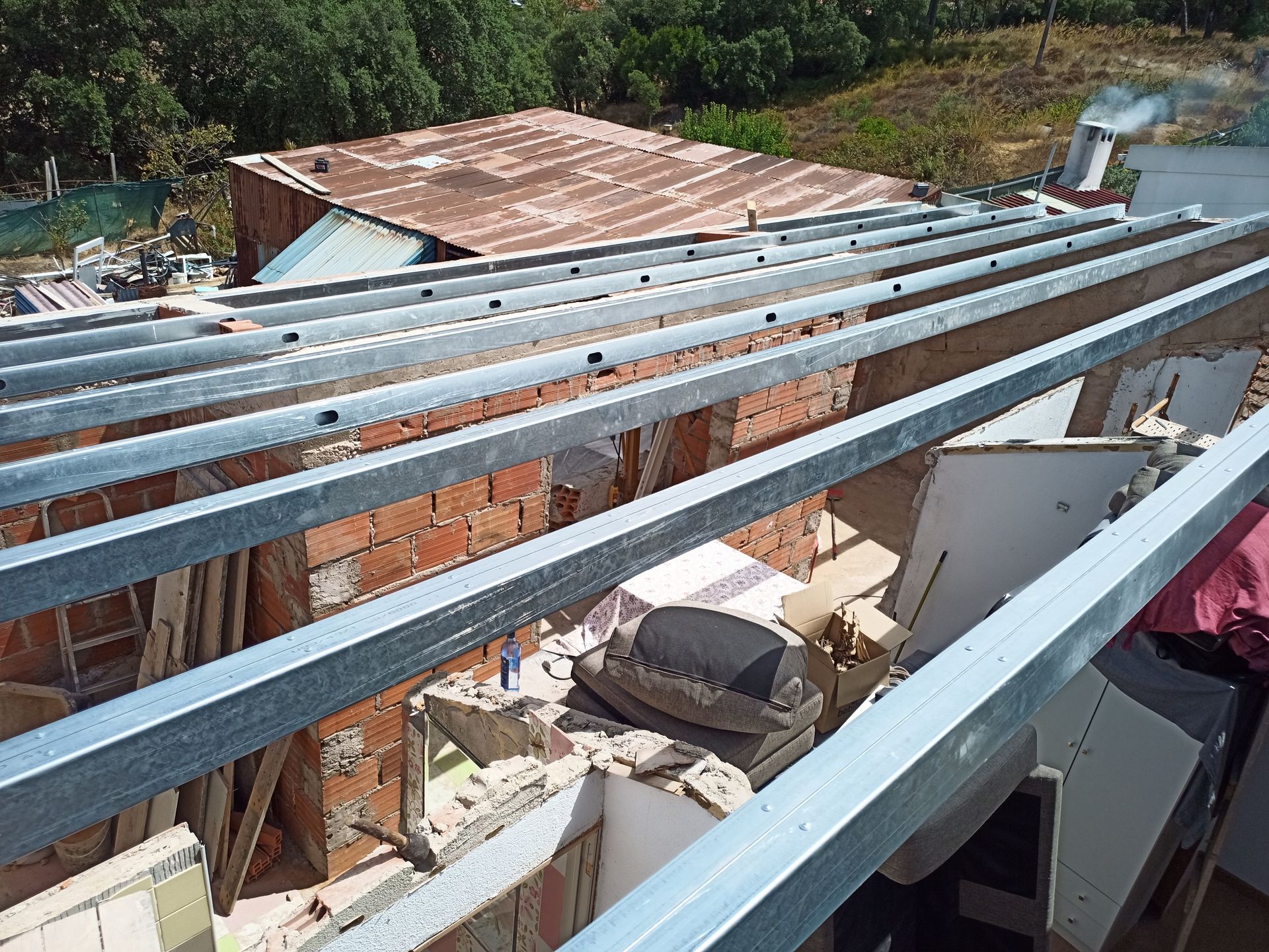 Canteiro de obras: Vigas metálicas em um prédio de tijolos parcialmente demolido, entulho visível e um telhado velho e enferrujado ao fundo.