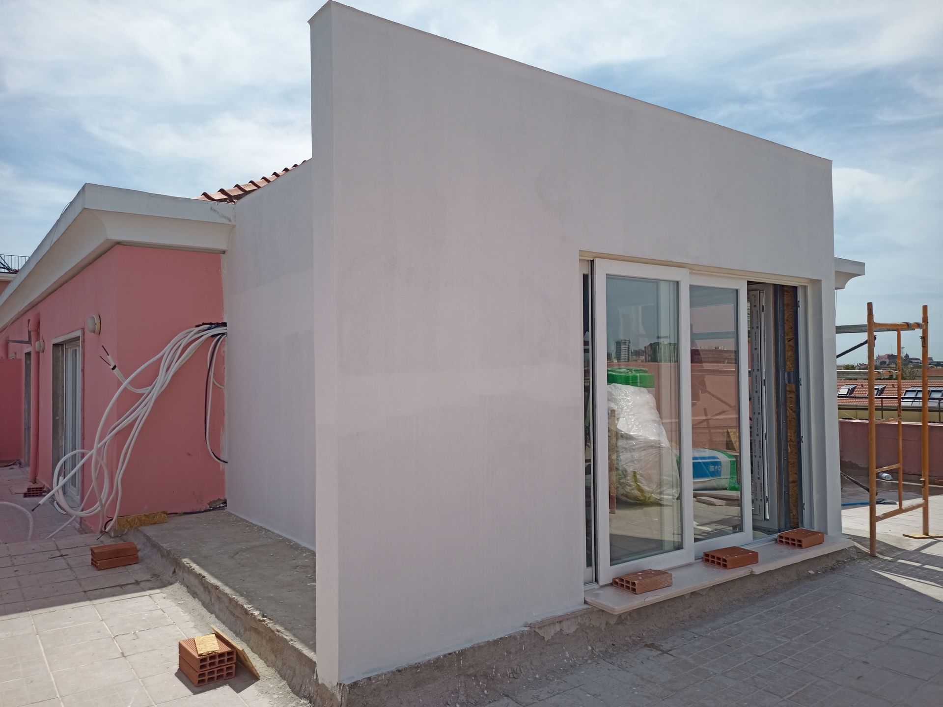 Exterior de um prédio em construção. Paredes rosa e brancas, porta de vidro deslizante, andaimes e céu ensolarado.