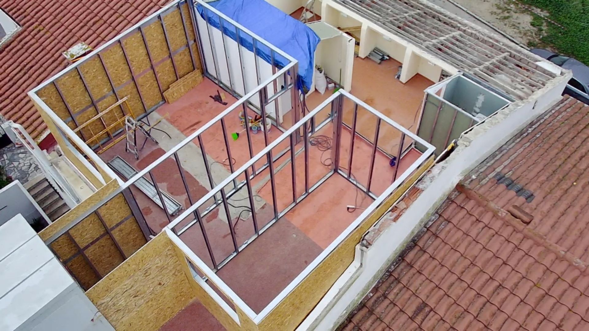 Vista aérea de um edifício em construção, mostrando paredes de vidro e madeira com uma lona azul cobrindo parte do telhado.