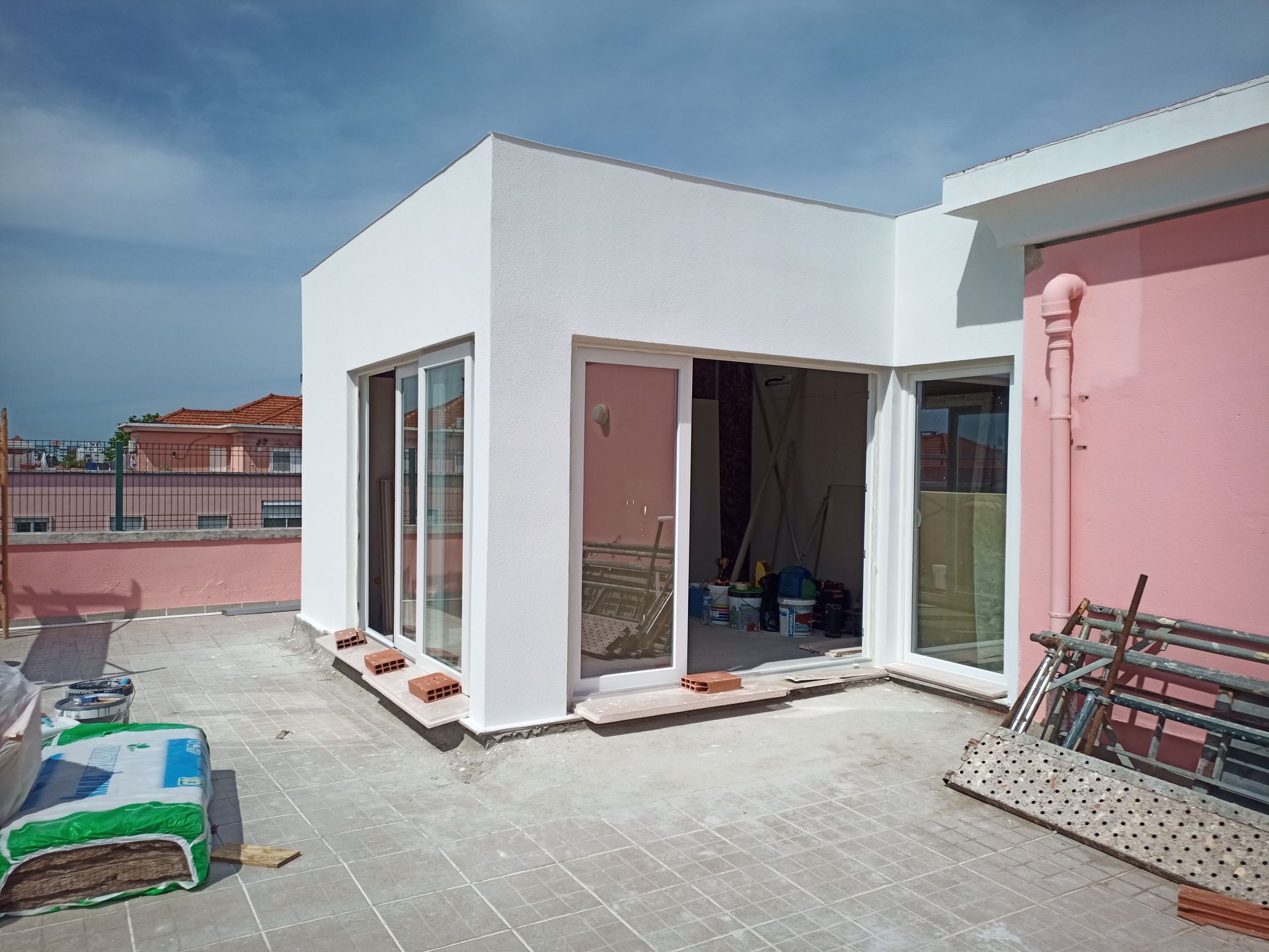 Construção no telhado: Estrutura com paredes brancas e portas de vidro deslizantes, interior inacabado, parede rosa e materiais de construção.