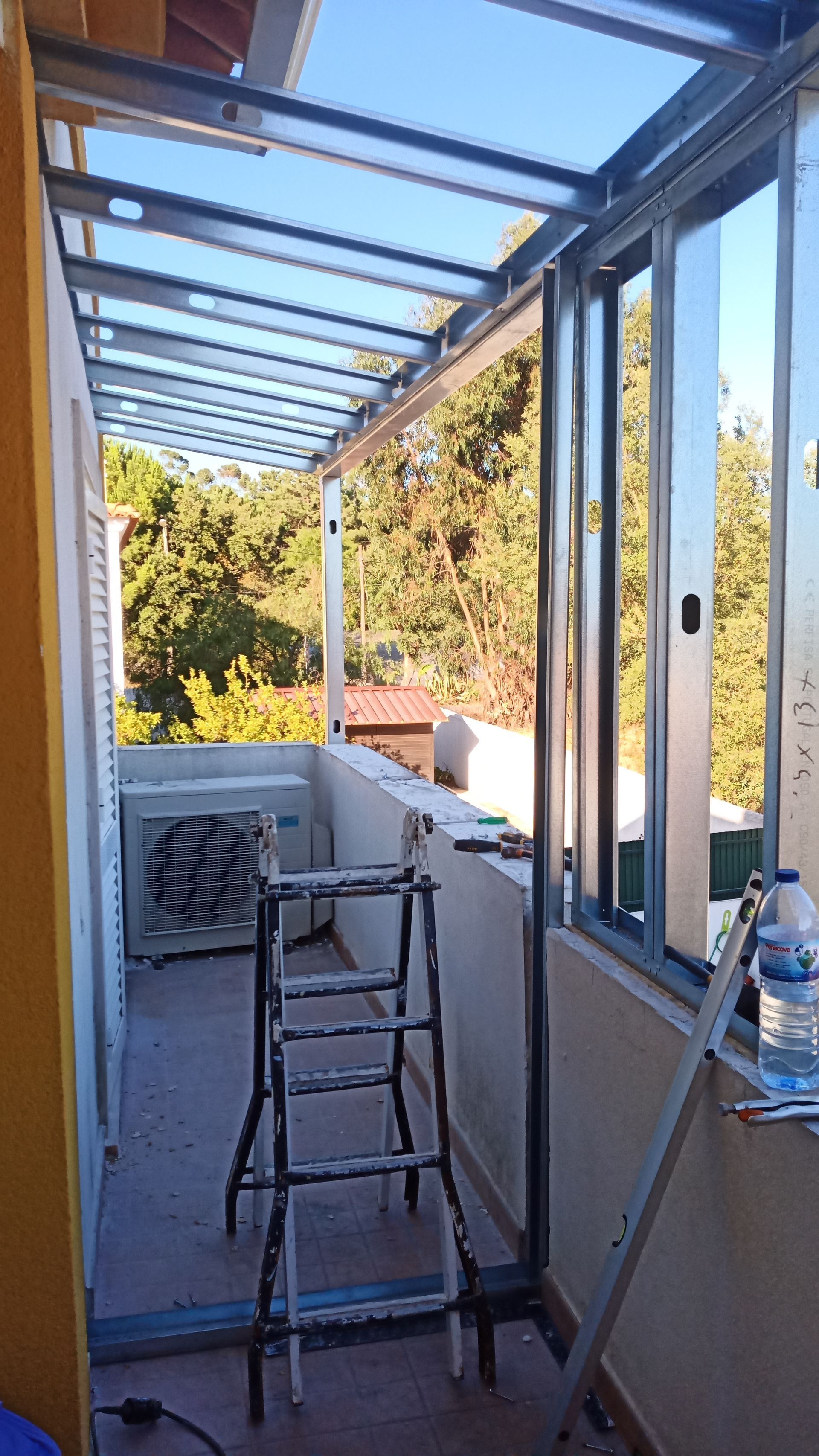 Varanda com estrutura metálica, painéis de cobertura transparentes, ar condicionado e escada. A luz do sol ilumina as árvores ao fundo.
