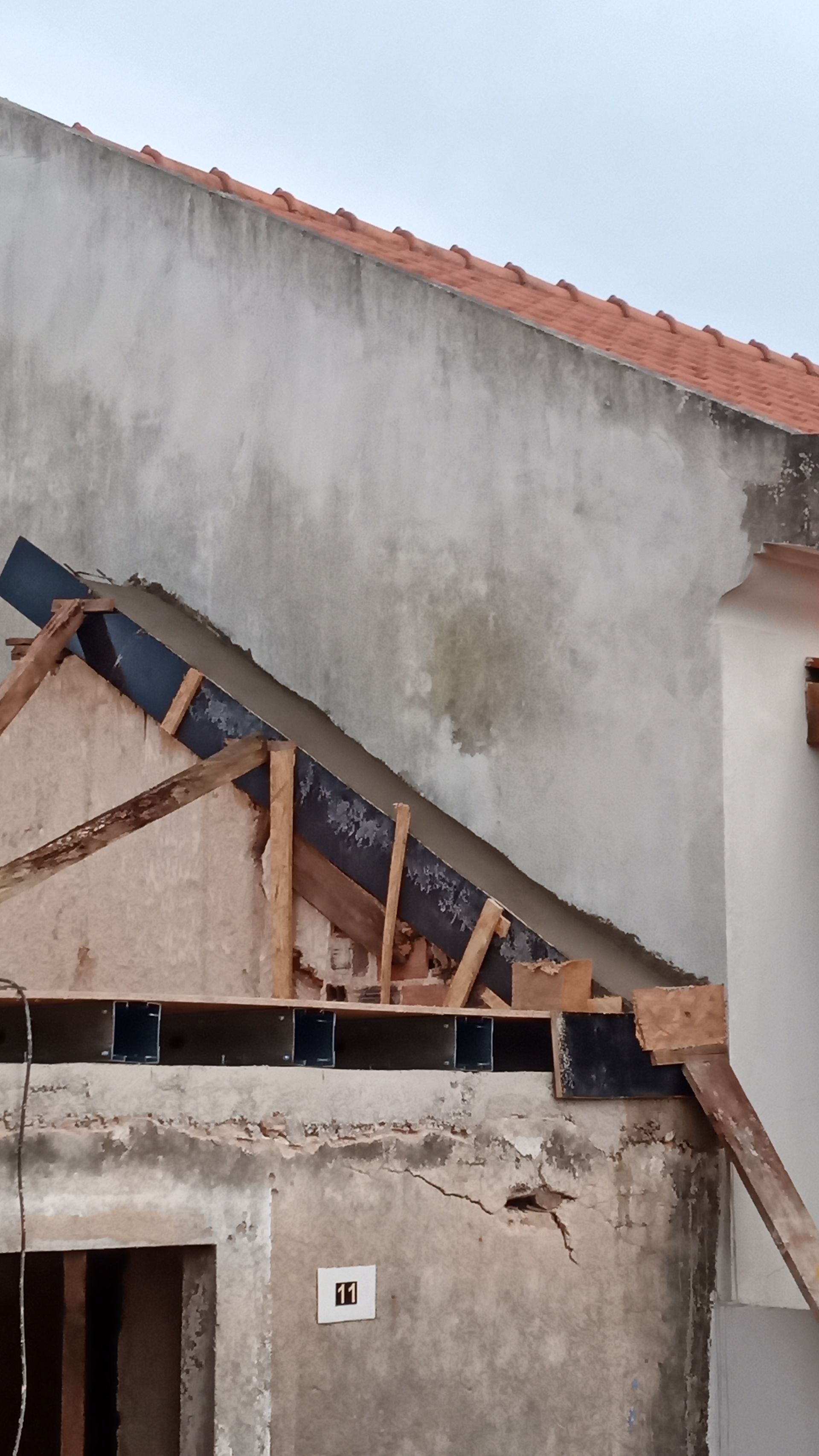 Fachada parcialmente construída de um edifício com vigas de madeira expostas, paredes cinzentas e telhado de telha vermelha.