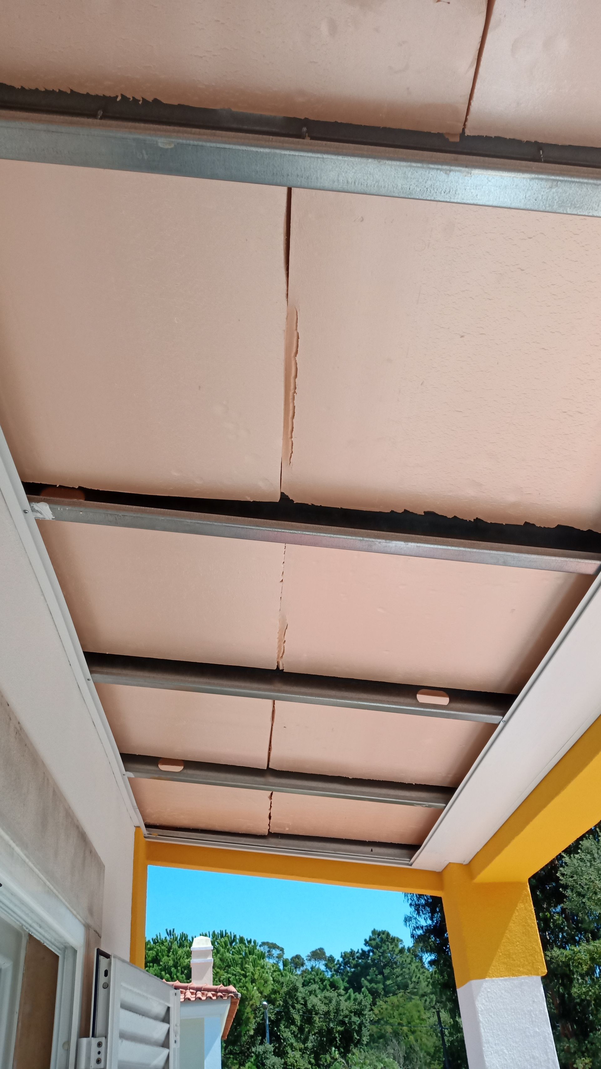 Painéis de teto bege instalados em estrutura metálica, com vista para o céu e as árvores abaixo.