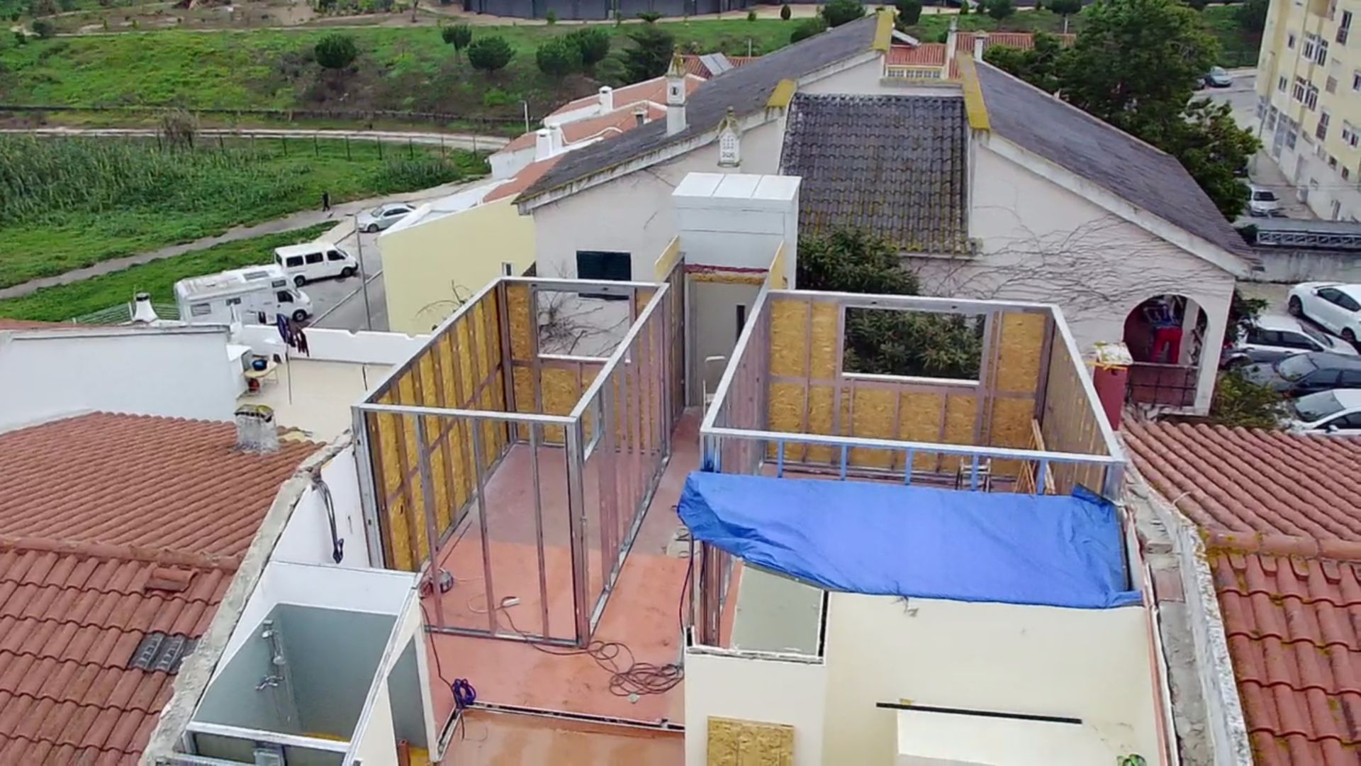 Construção da estrutura do telhado de um edifício, mostrando a estrutura aparente e a lona azul, cercada por outros edifícios.