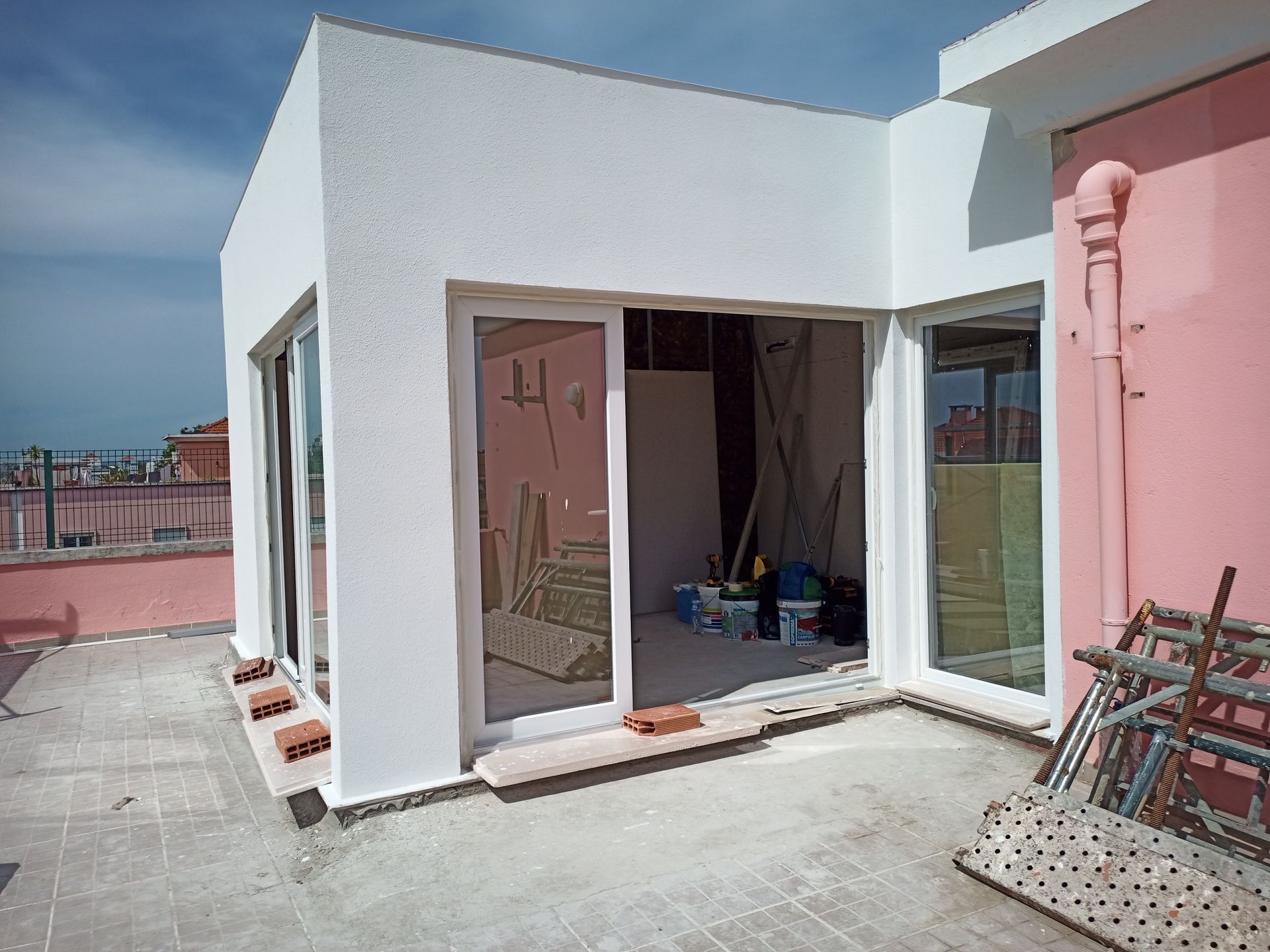 Vista exterior de um edifício pintado de branco com portas de vidro deslizantes, interior inacabado e parede rosa na lateral.