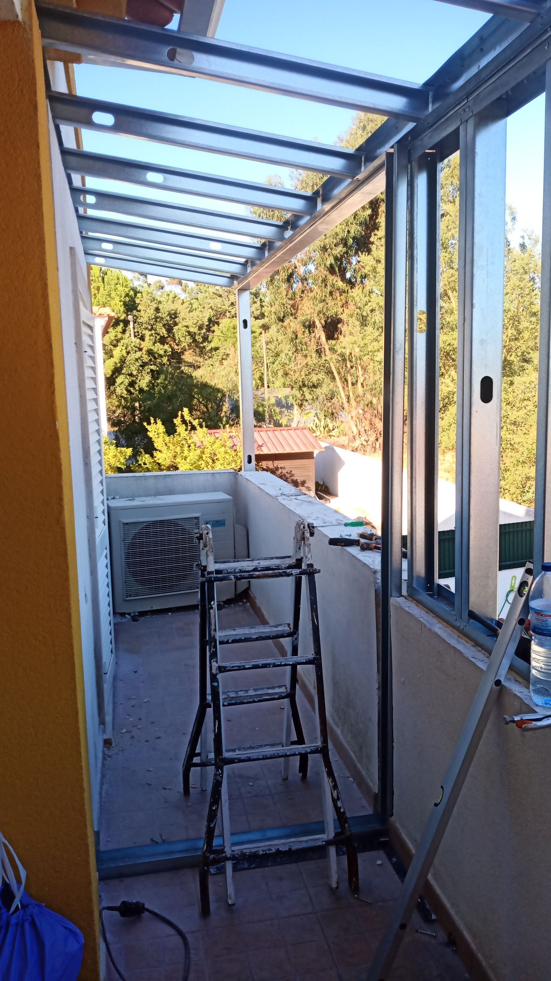 Varanda em construção com estrutura metálica, escada e ar condicionado. Dia ensolarado, árvores ao fundo.