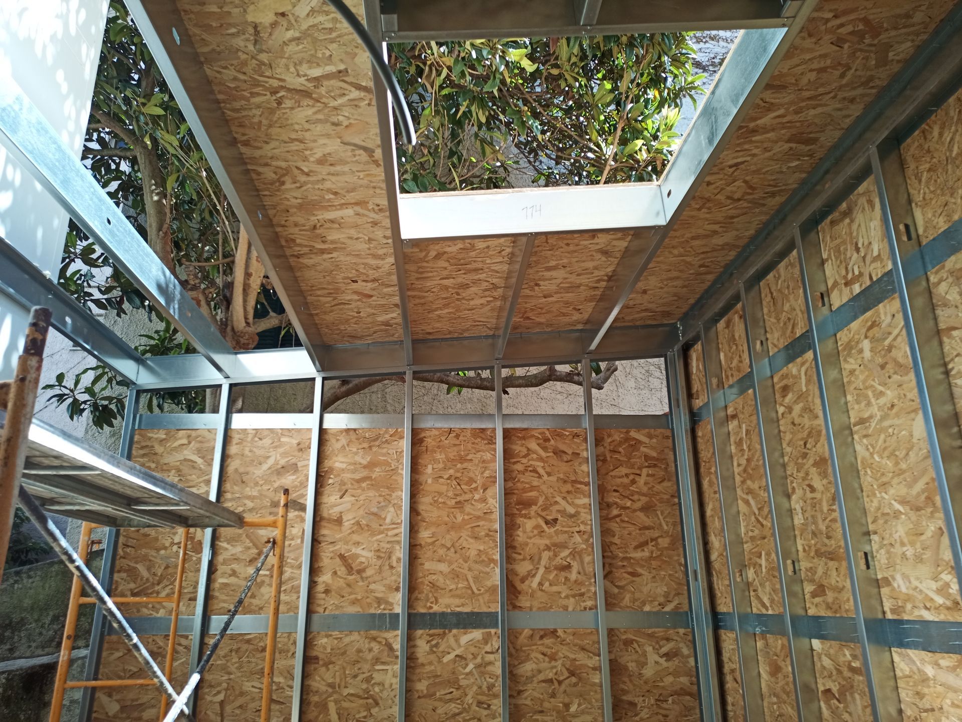 Vista interna de um cômodo em construção com estrutura metálica, paredes de painéis OSB e uma claraboia.