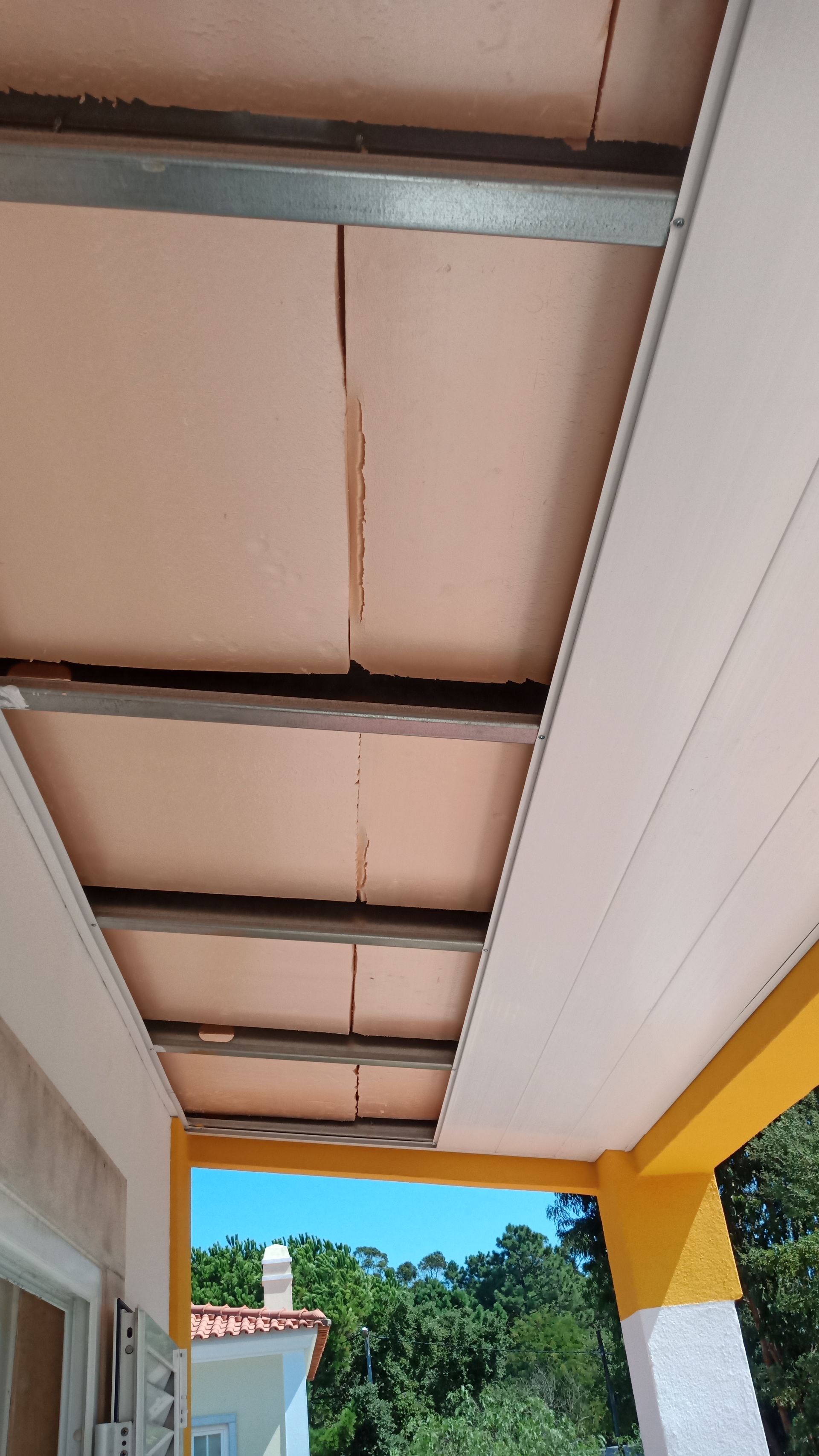 Estrutura do teto: painéis de isolamento rosa entre suportes metálicos, adjacentes a painéis brancos, colunas pintadas de amarelo e branco.