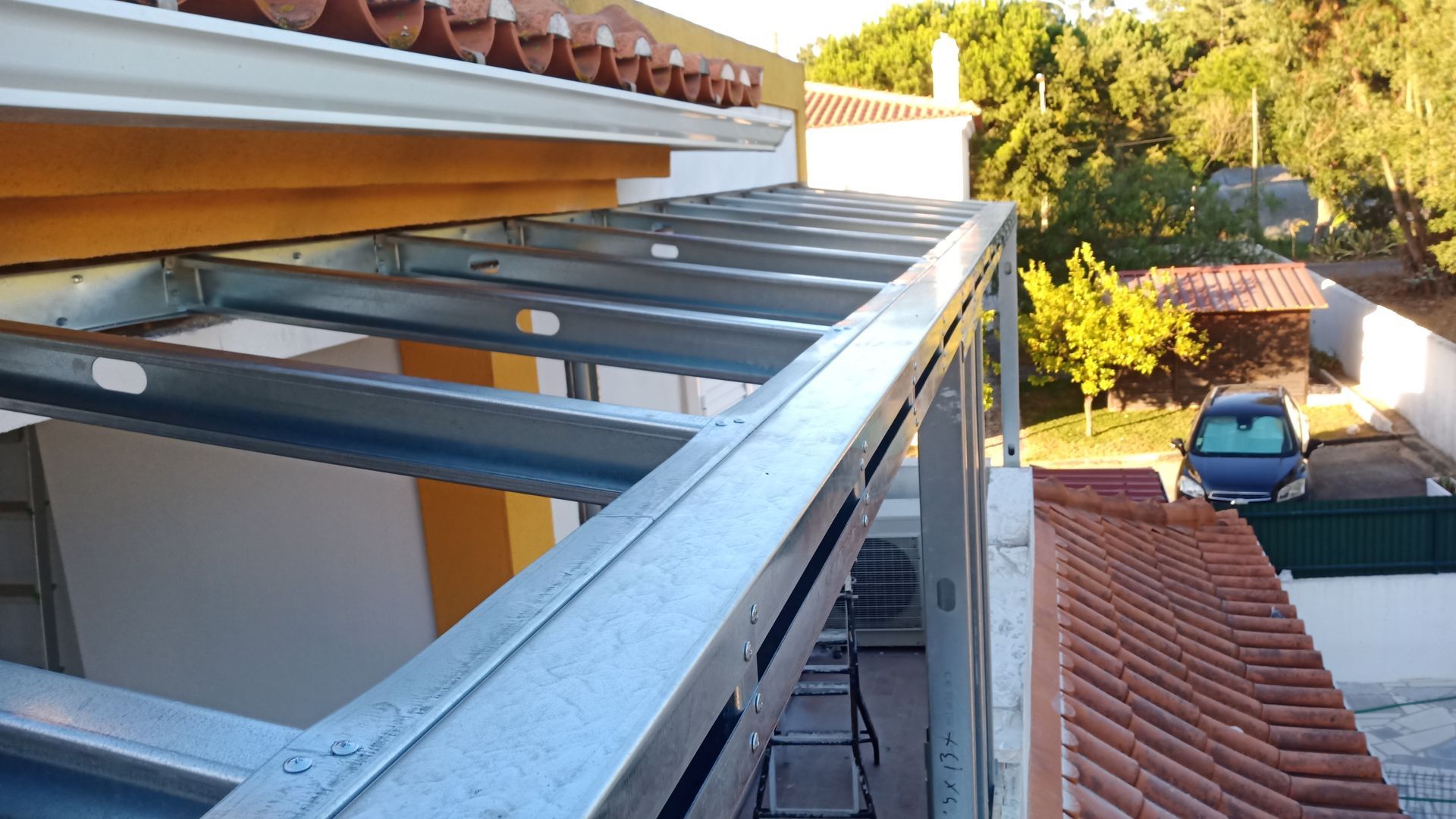 Cobertura com estrutura metálica fixada a um edifício, pronta para receber o telhado, ao lado de um telhado de telhas vermelhas e vegetação.