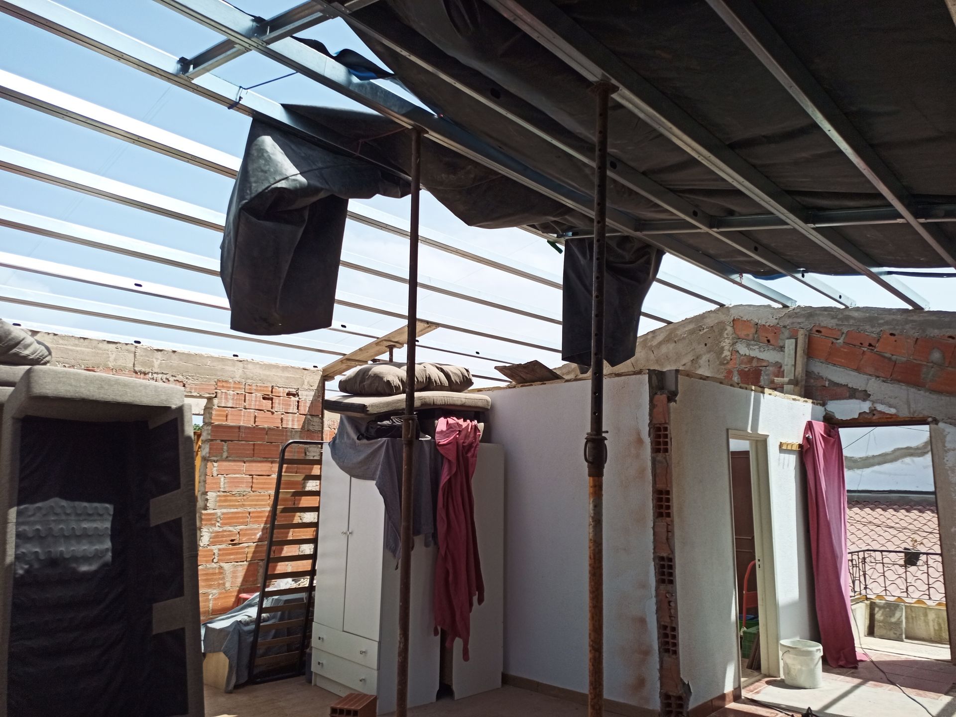 Canteiro de obras com paredes de tijolos aparentes, teto com estrutura metálica e postes de sustentação. Lonas estão estendidas sobre a estrutura.