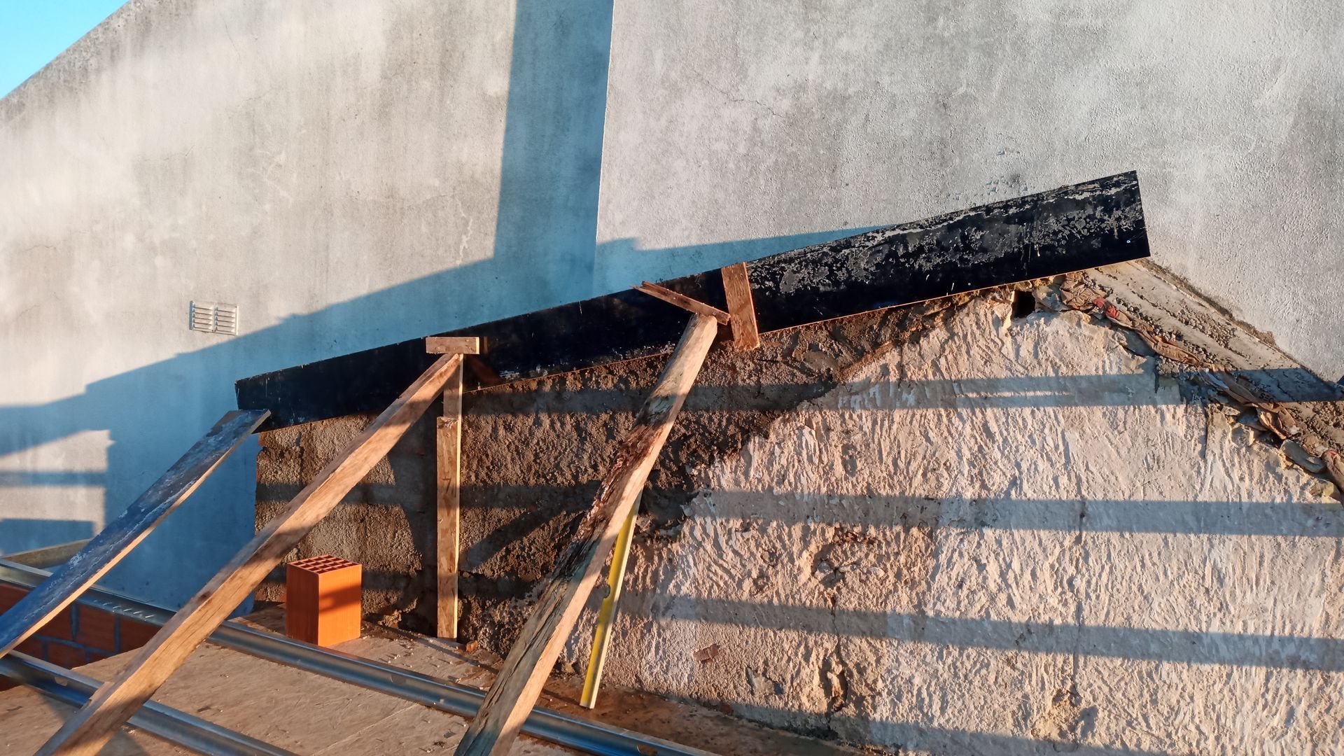 Parede com parte do telhado em construção; suportes de madeira, fita de vedação preta e tijolo.