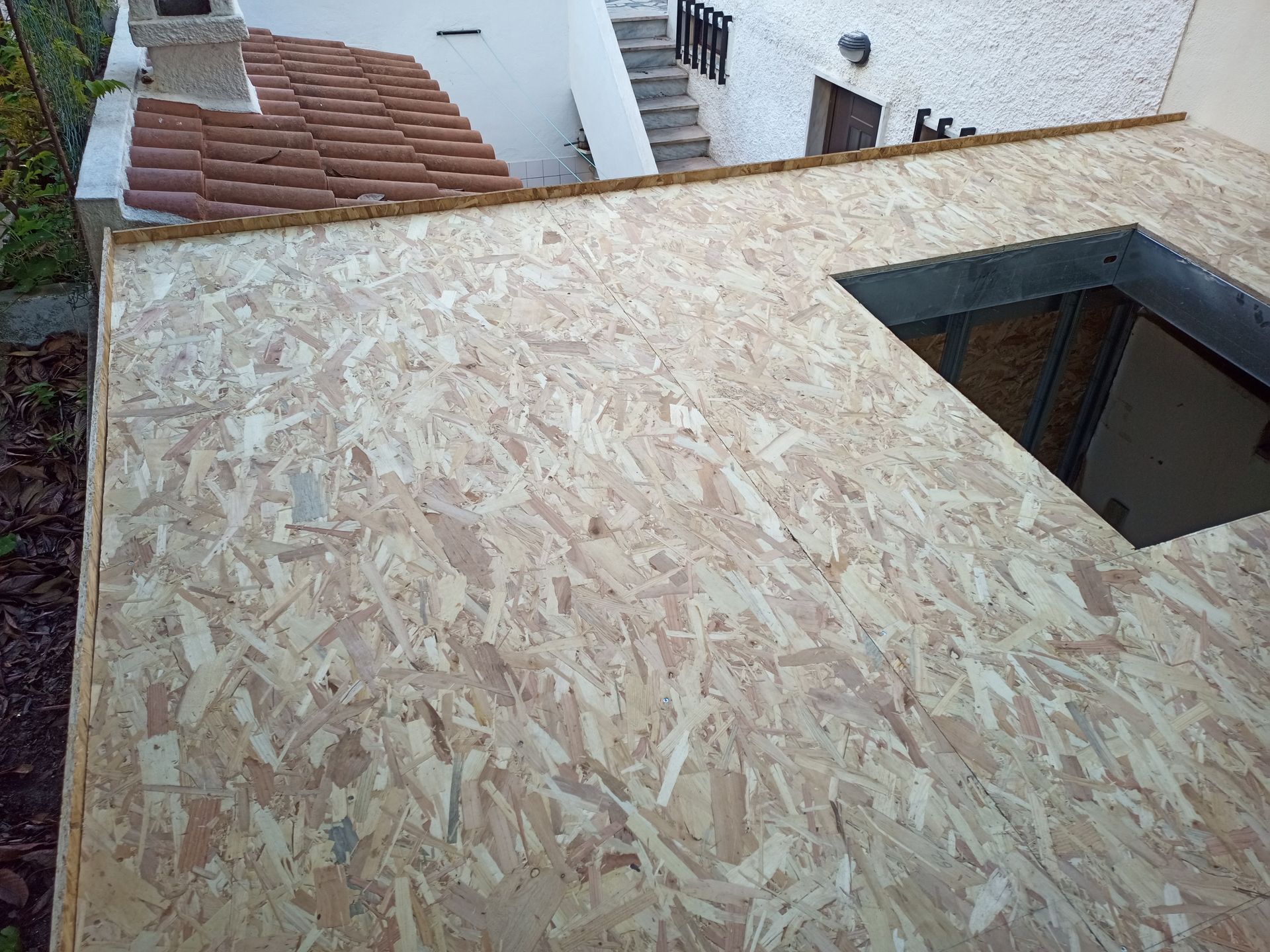 Telhado de OSB com abertura quadrada, adjacente a um telhado de telha e um edifício branco.