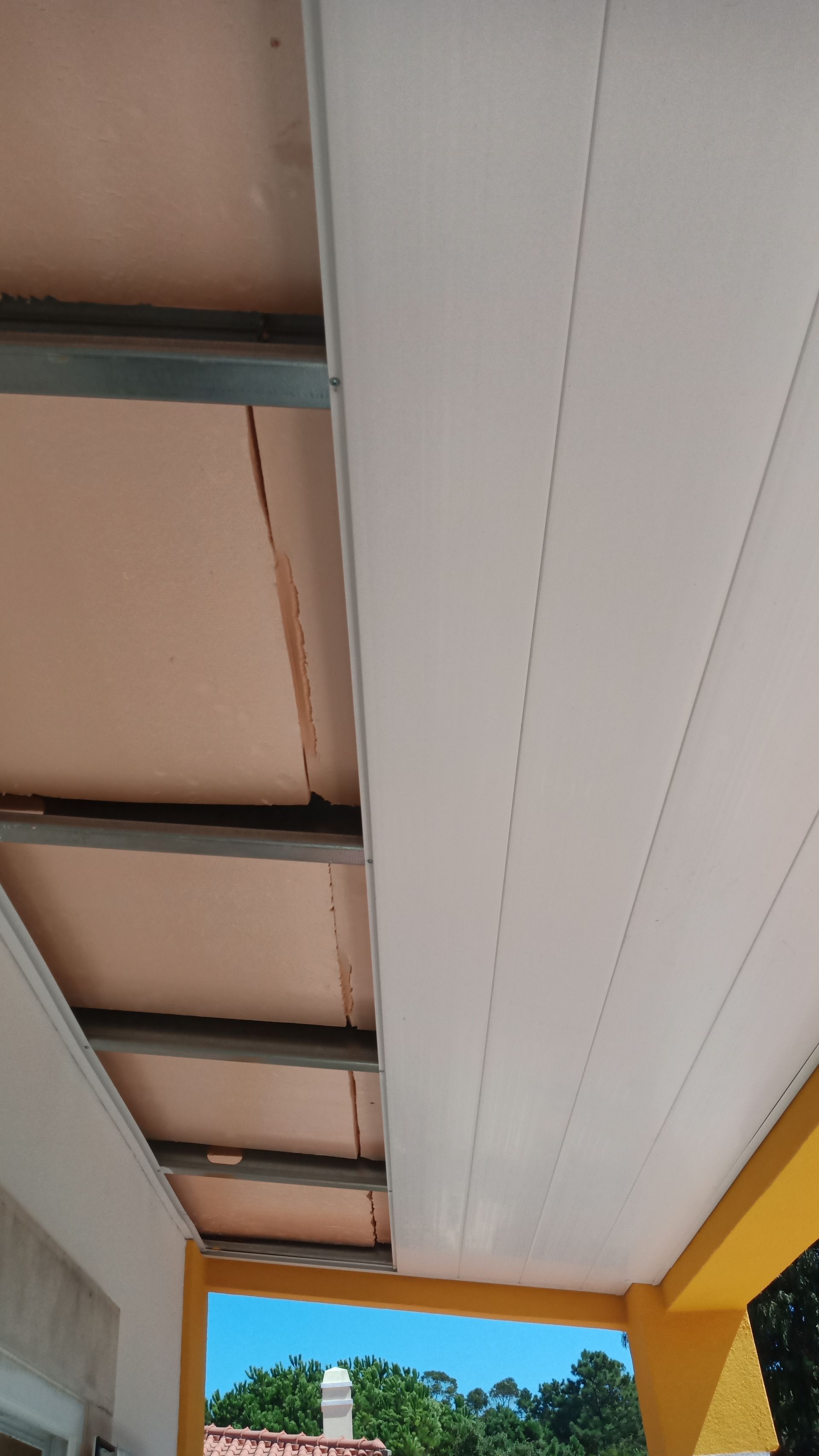 Painéis de teto brancos sob uma estrutura metálica. Isolamento rosa acima. Exterior do edifício em amarelo e branco.