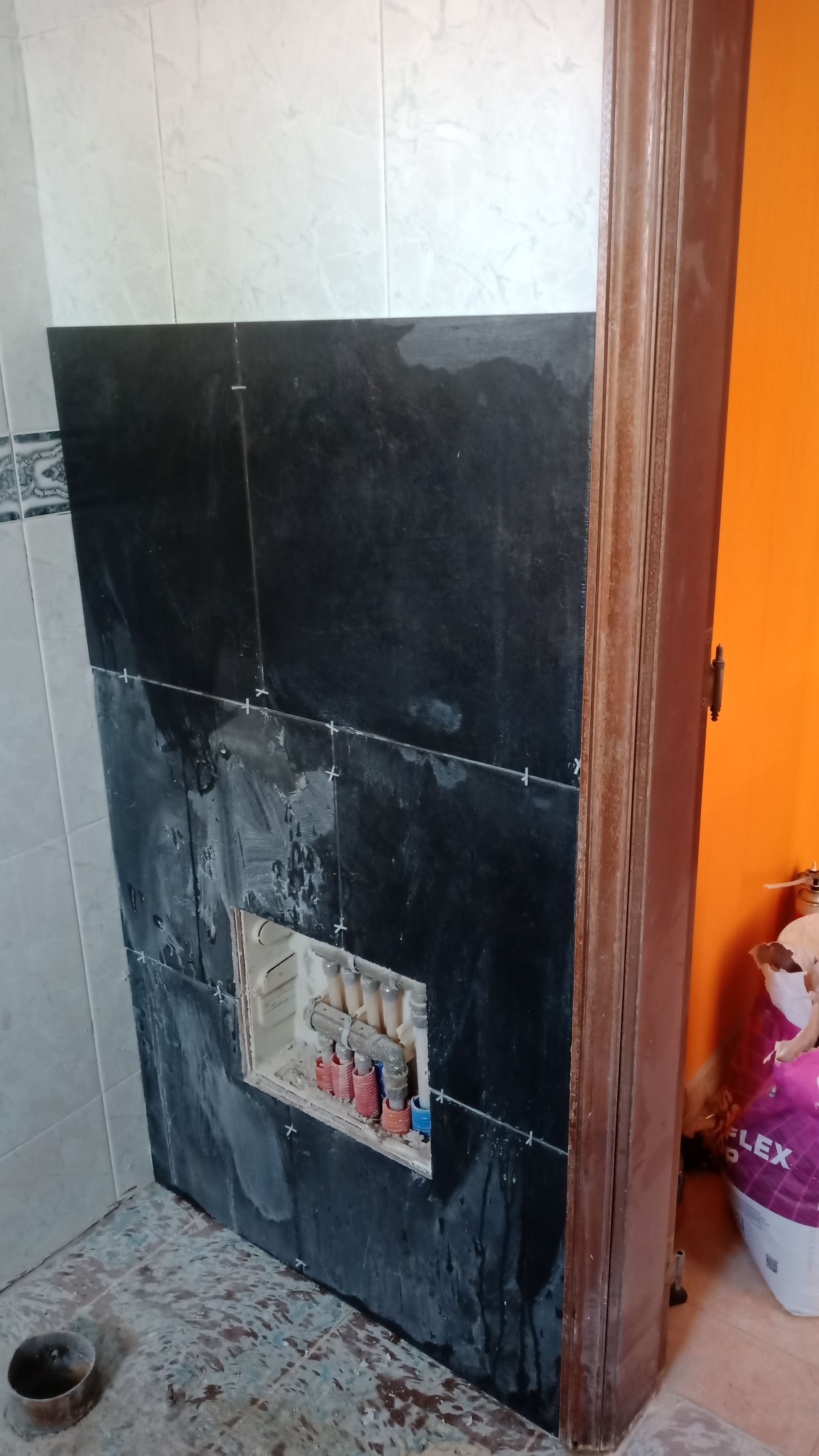 Parede revestida com azulejos pretos e com uma prateleira embutida, ao lado de uma moldura de porta marrom. Vista parcial de uma parede revestida com azulejos brancos.