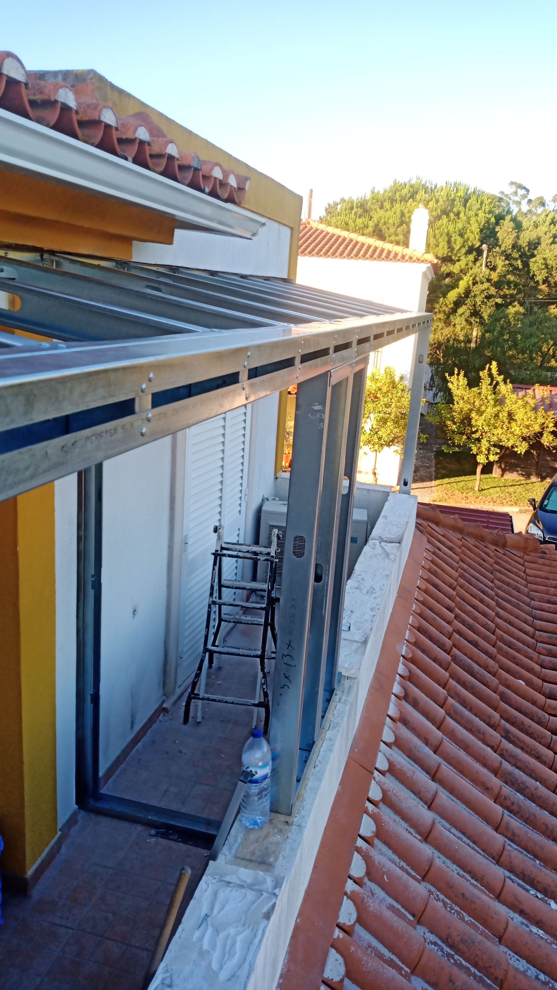Toldo em construção sobre uma varanda. Estrutura metálica instalada com escada, com vista para um telhado de terracota e árvores.