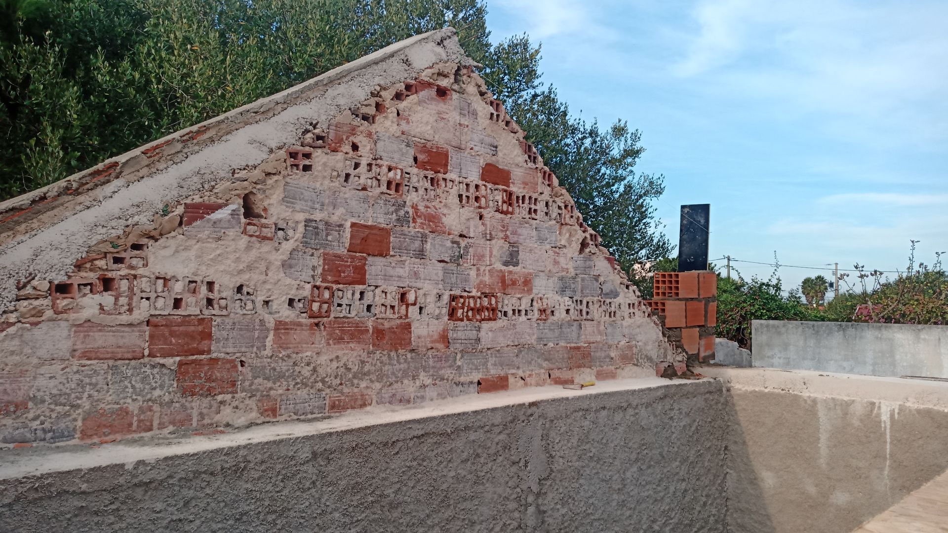 Estrutura de tijolos com argamassa deteriorada, parcialmente revestida de estuque, ao lado de uma superfície de concreto, contra um céu azul.