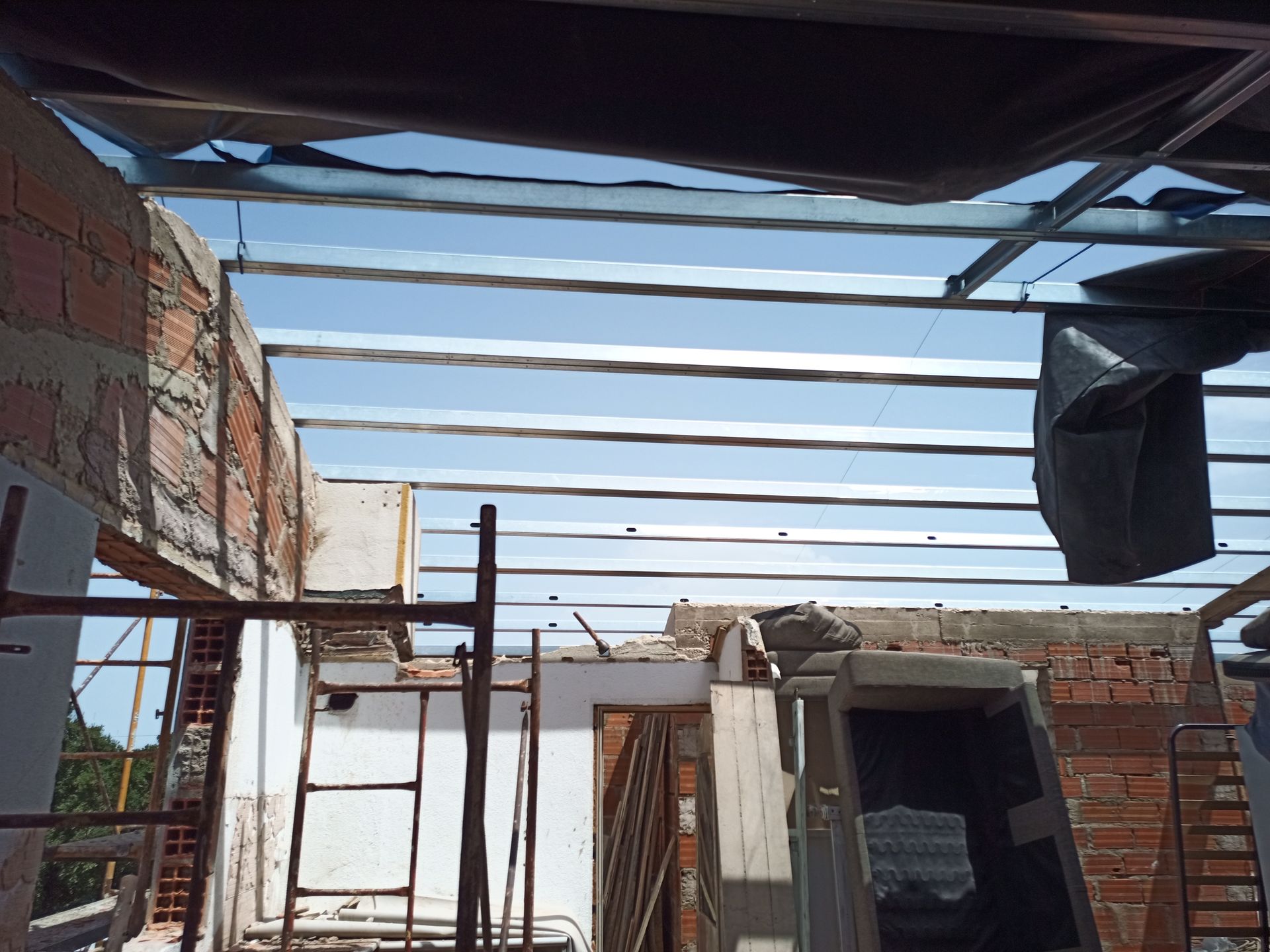 Canteiro de obras com vigas metálicas expostas, paredes de tijolos e vista para o céu.