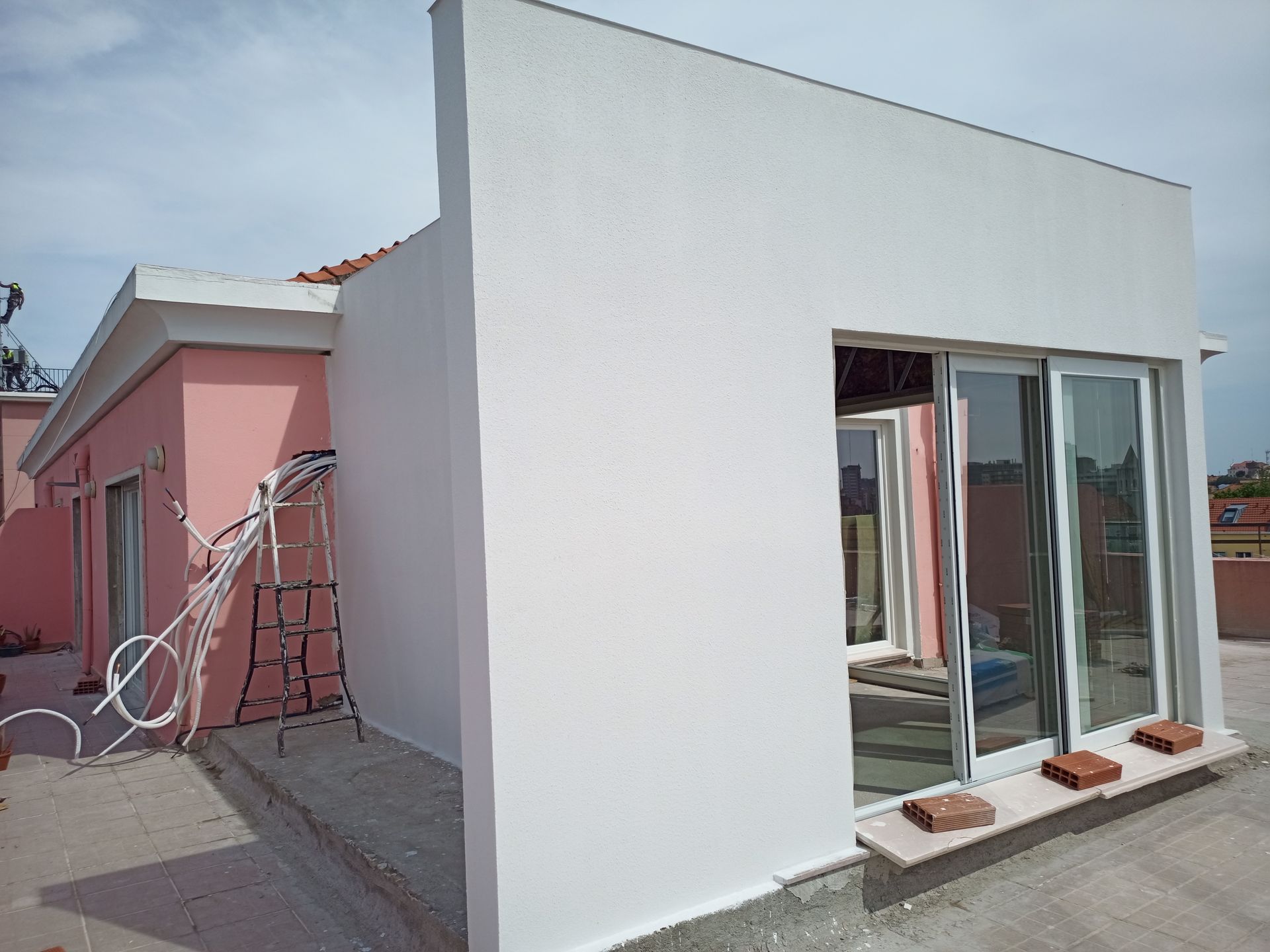 Exterior de um edifício em construção, com paredes rosa e brancas, uma porta de vidro deslizante e fios elétricos expostos.