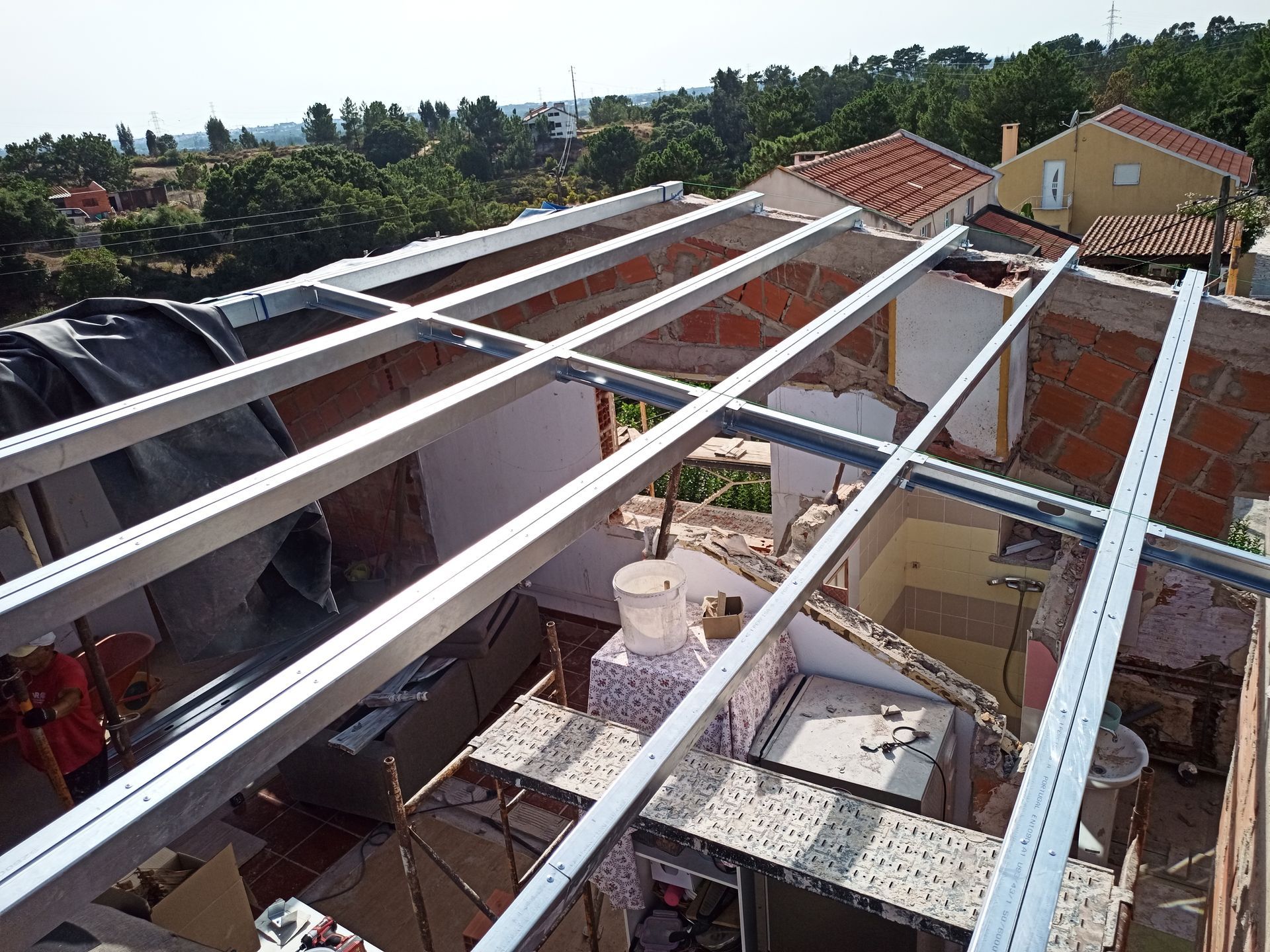 Vista aérea de uma casa em fase de construção do telhado; vigas metálicas atravessam o interior exposto, com paredes de tijolos e árvores ao redor visíveis.