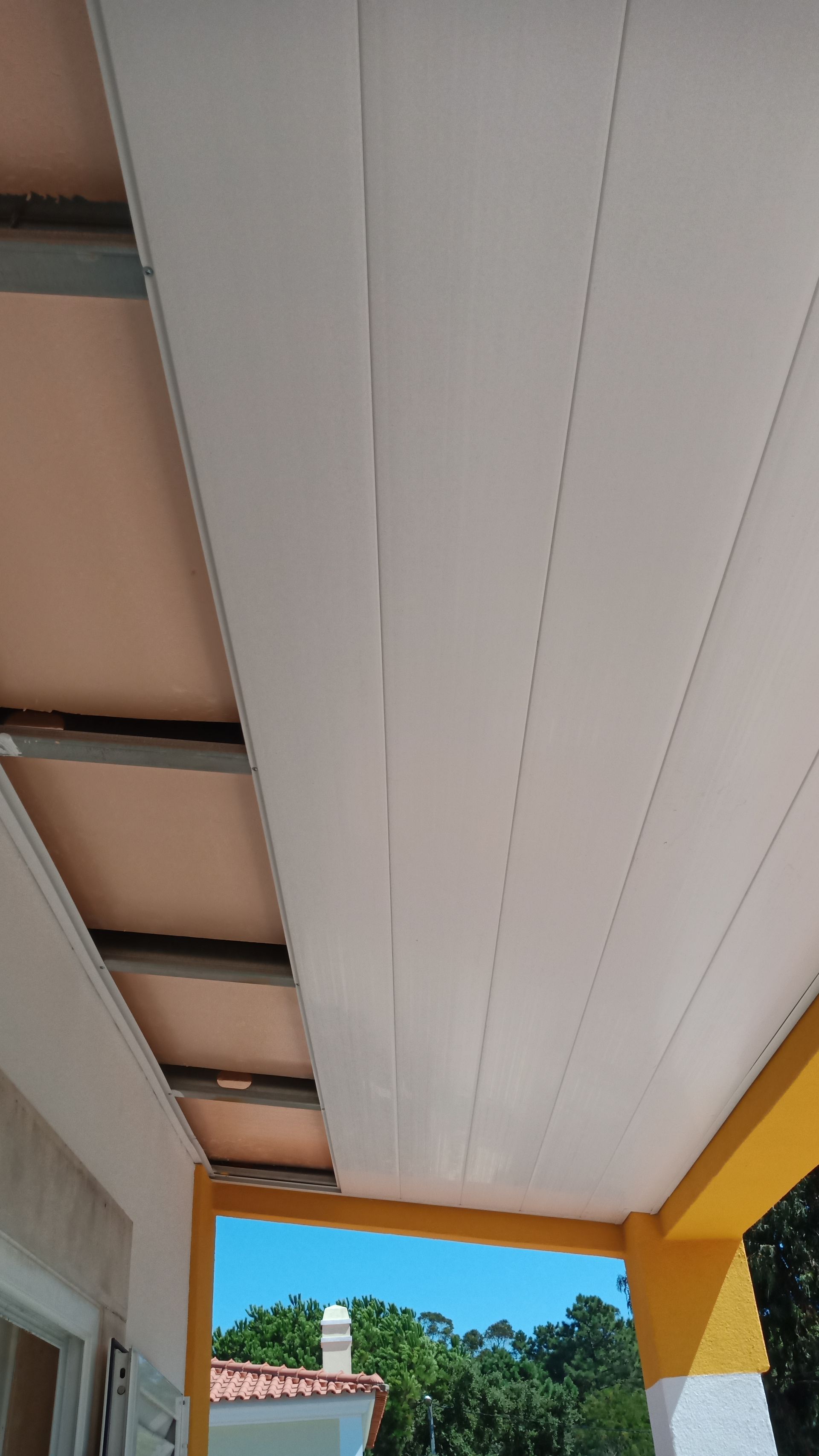 Teto com painéis brancos e vigas de sustentação visíveis, visto de baixo para cima a partir de uma varanda amarela.