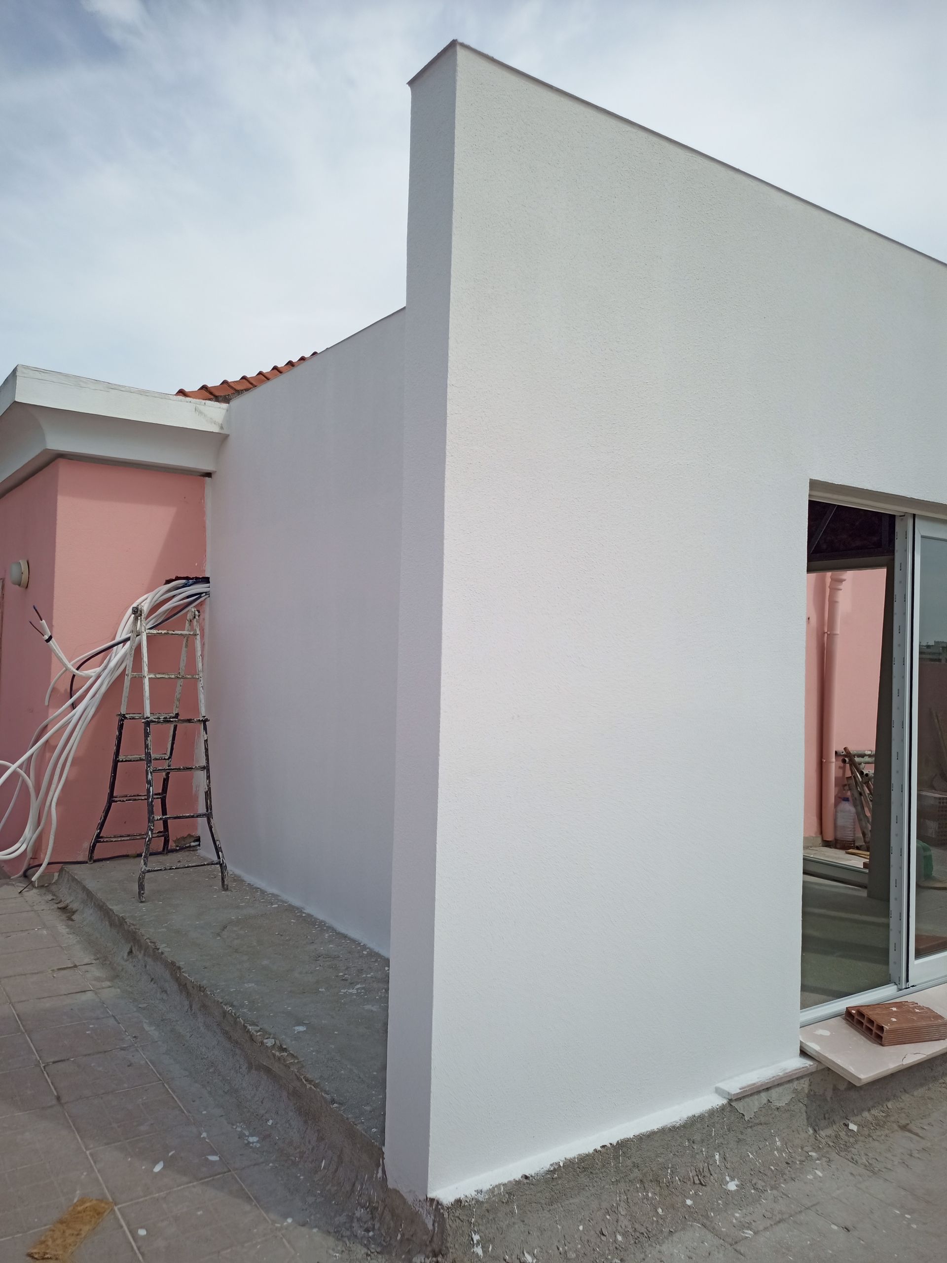 Fachada externa de estuque branco de um prédio com uma janela, ao lado de uma parede rosa com fiação elétrica. Céu nublado.