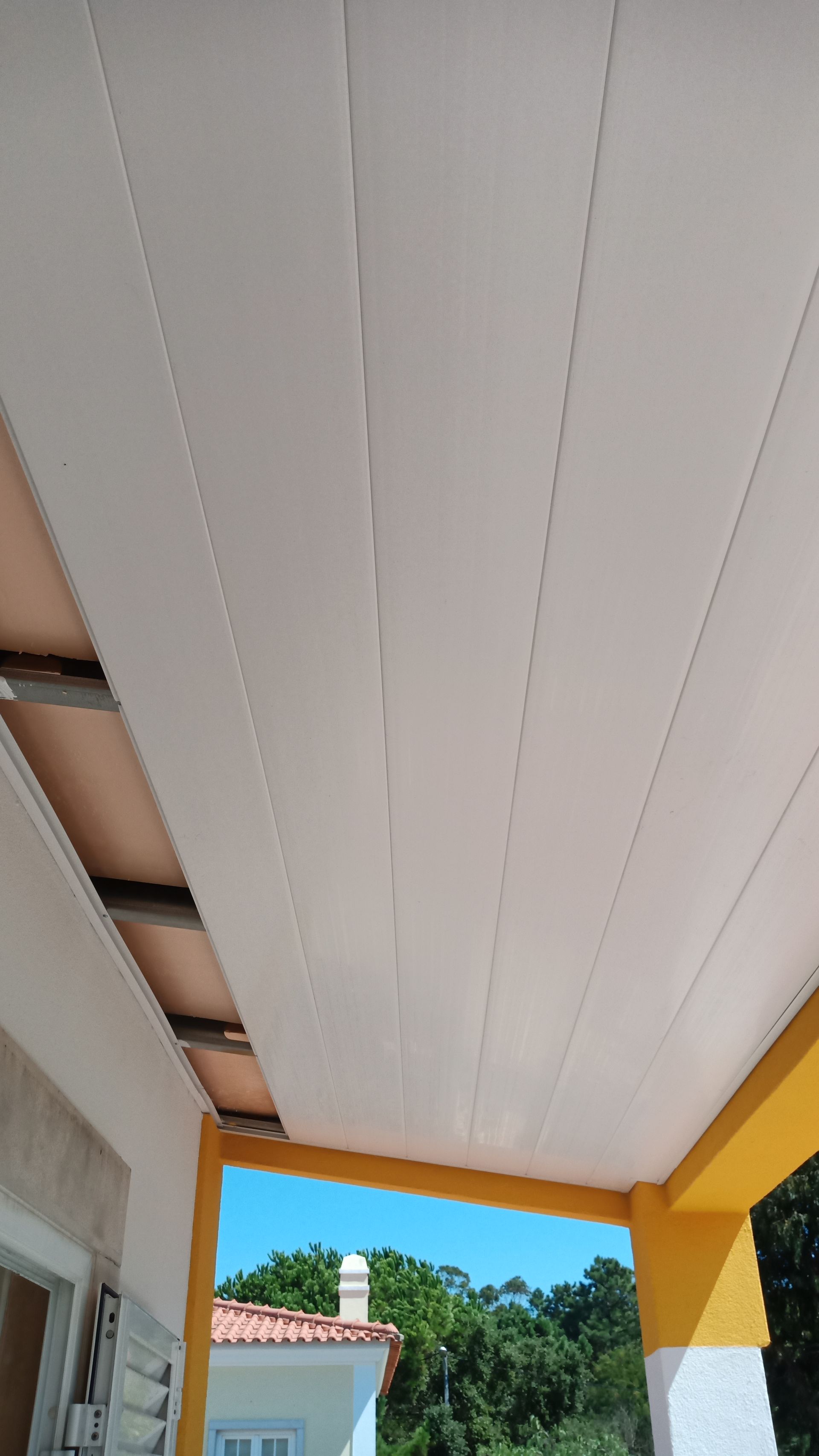 Teto com painéis brancos e vista para o céu azul, árvores e um prédio com detalhes em laranja e amarelo.
