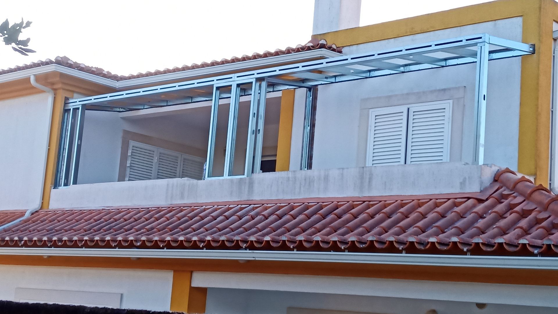Estrutura metálica de varanda em um prédio amarelo com telhado de telha vermelha.