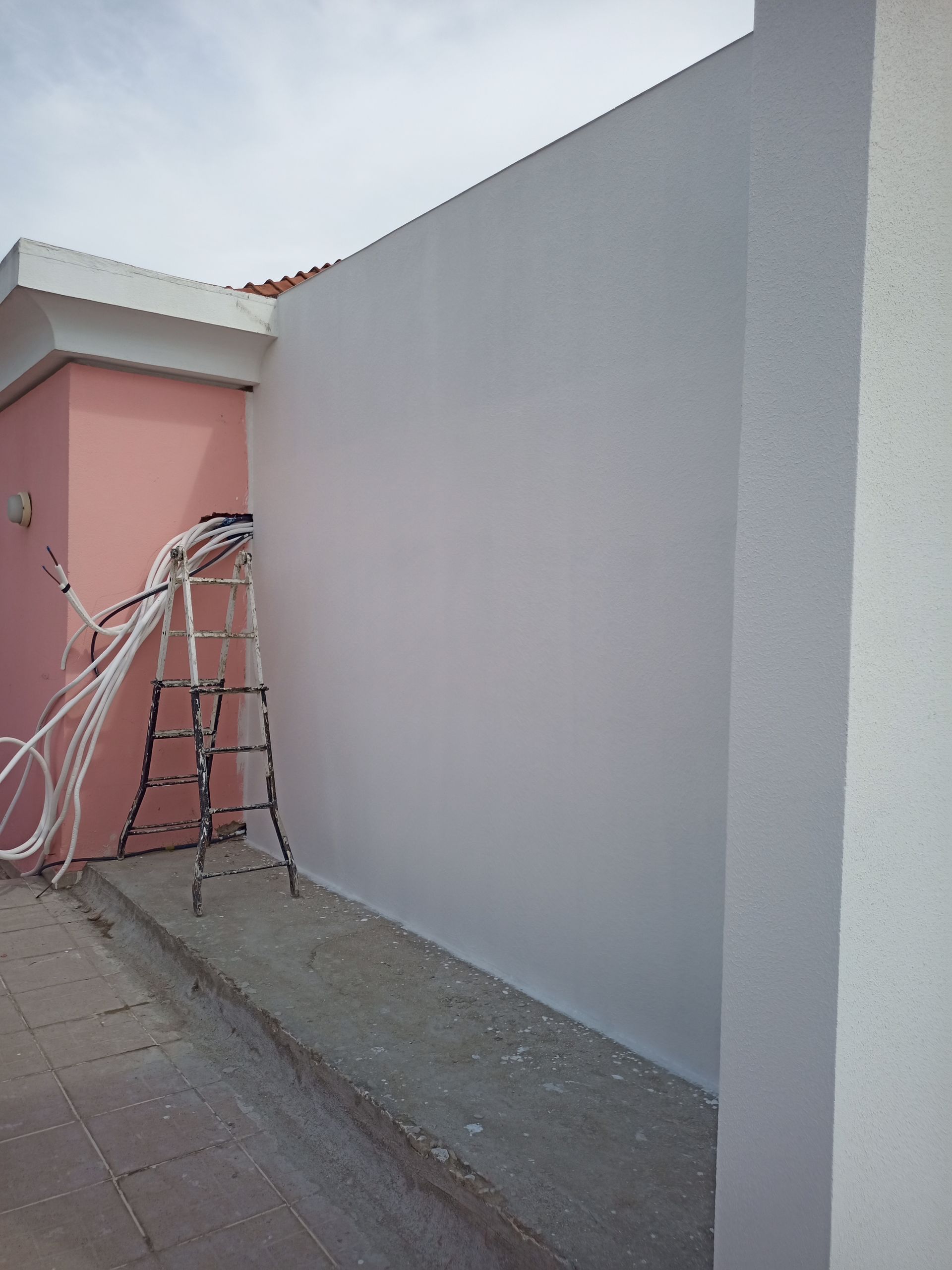 Parede pintada de cinza adjacente a um prédio rosa, com escada e fios presentes.