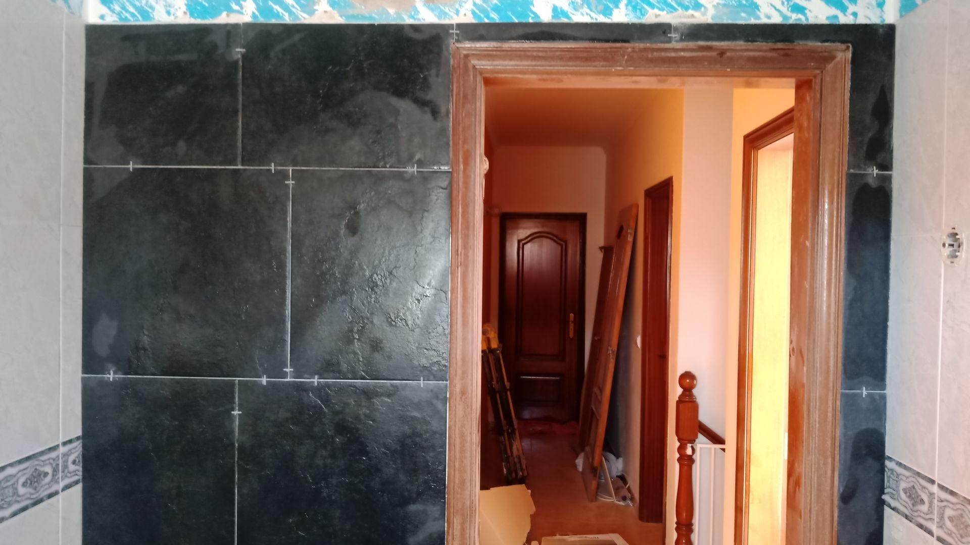 Parede revestida com azulejos escuros e porta que dá acesso a um corredor com portas e corrimãos de madeira.