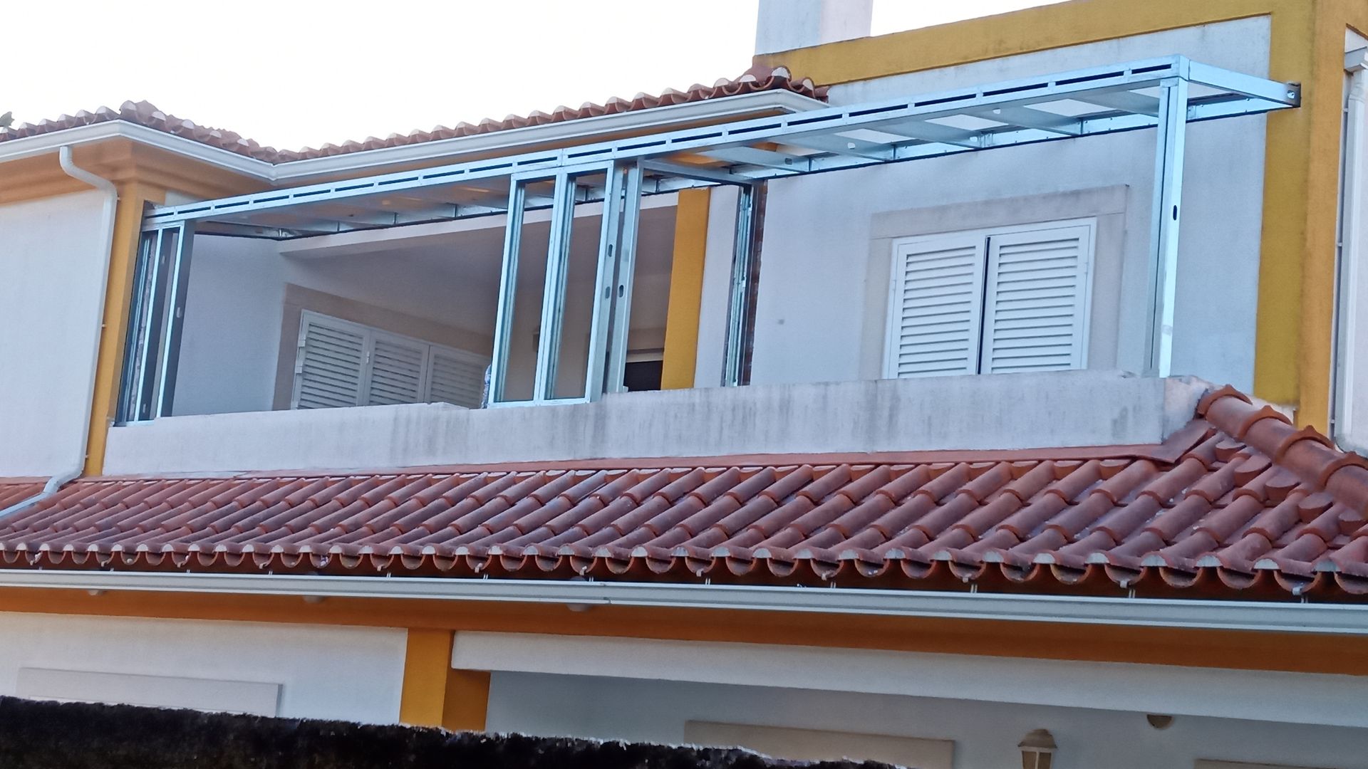 Toldo de metal sobre uma varanda, fixado em um prédio branco com telhado de telhas vermelhas.