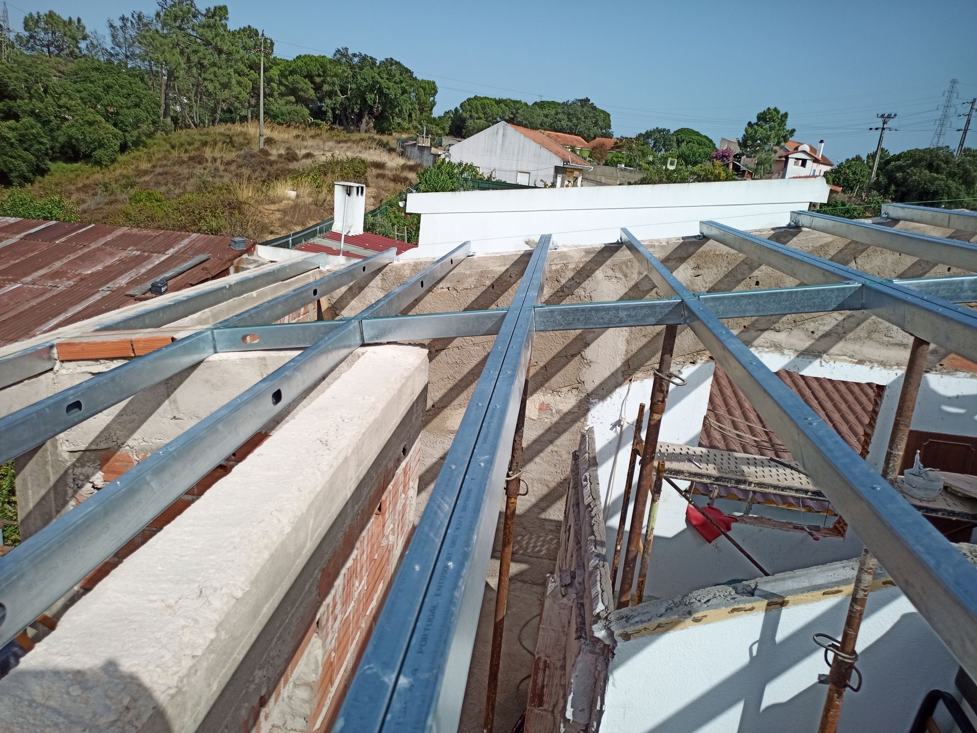 Estrutura metálica em um telhado em construção, com paredes de tijolos visíveis e edifícios circundantes sob um céu ensolarado.
