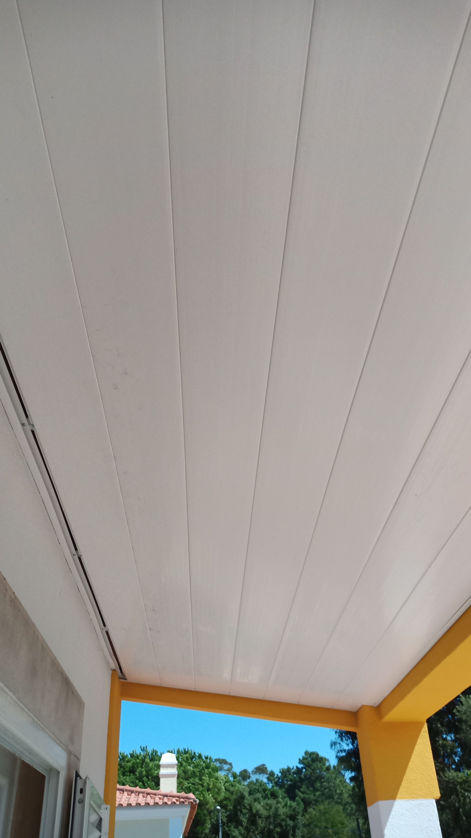 Teto com painéis brancos sobre uma varanda com detalhes em amarelo; céu azul e árvores visíveis abaixo.