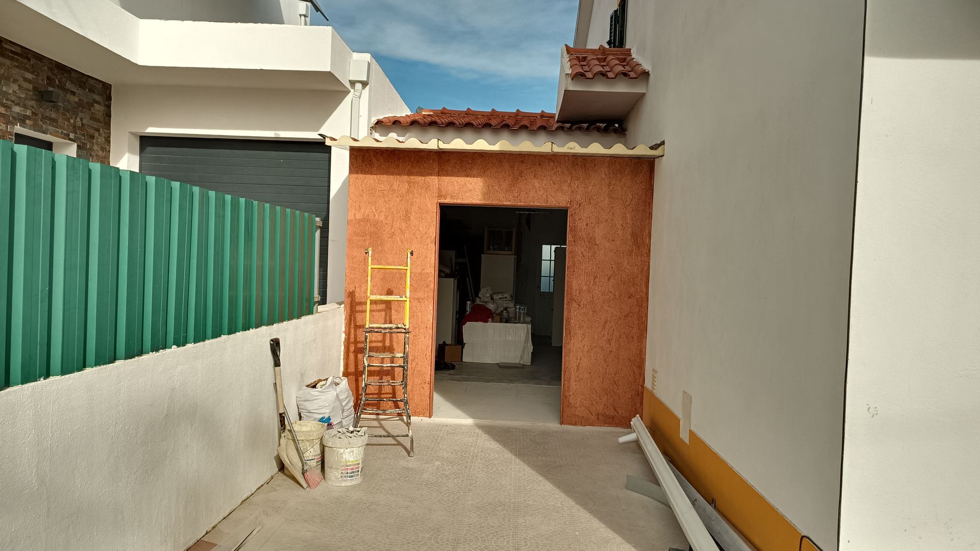 Beco estreito com obra em andamento; entrada com revestimento laranja, ferramentas e cerca verde.