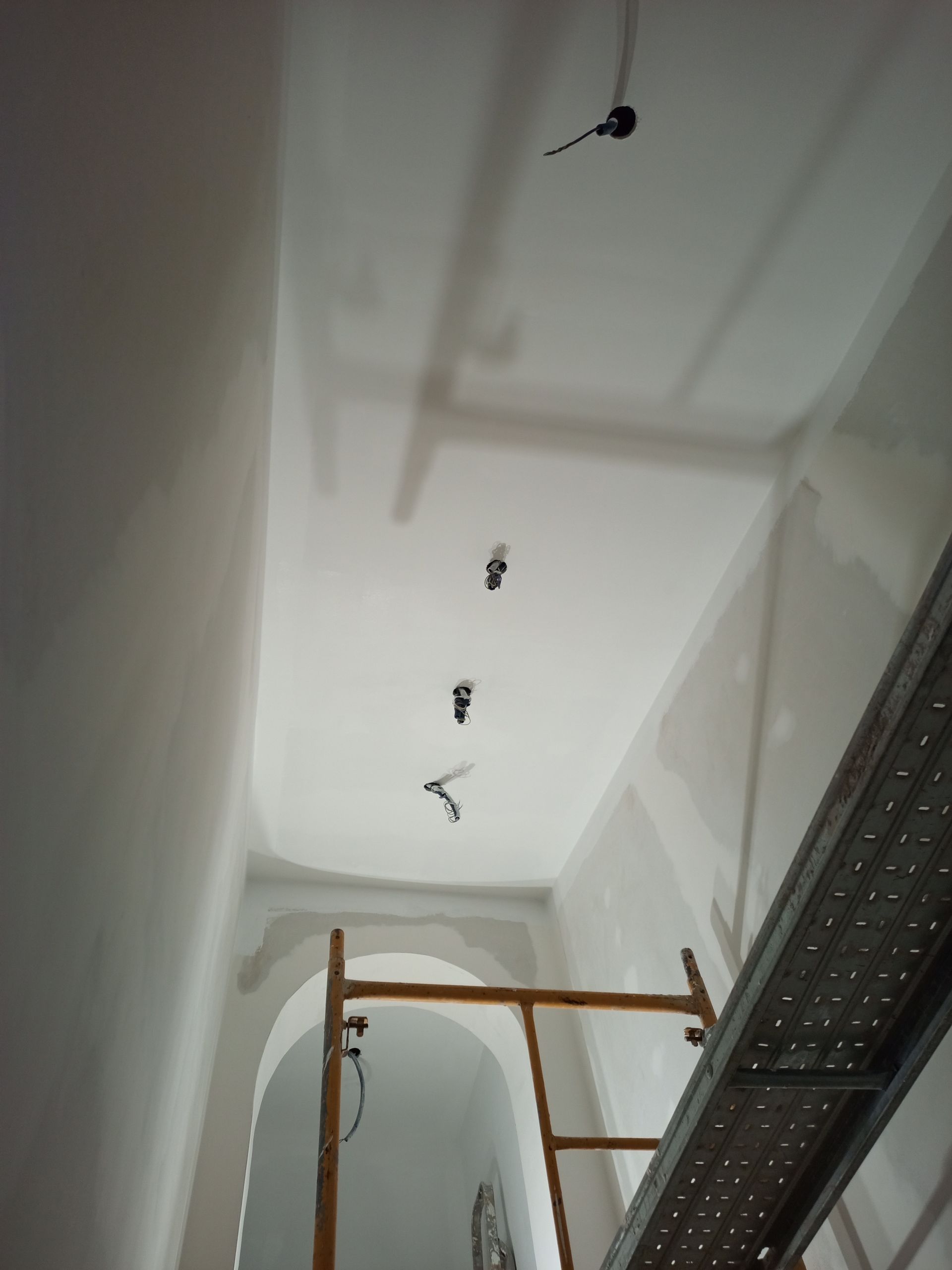 Vista interna de uma sala em construção com fiação exposta, andaimes e drywall no teto e nas paredes.