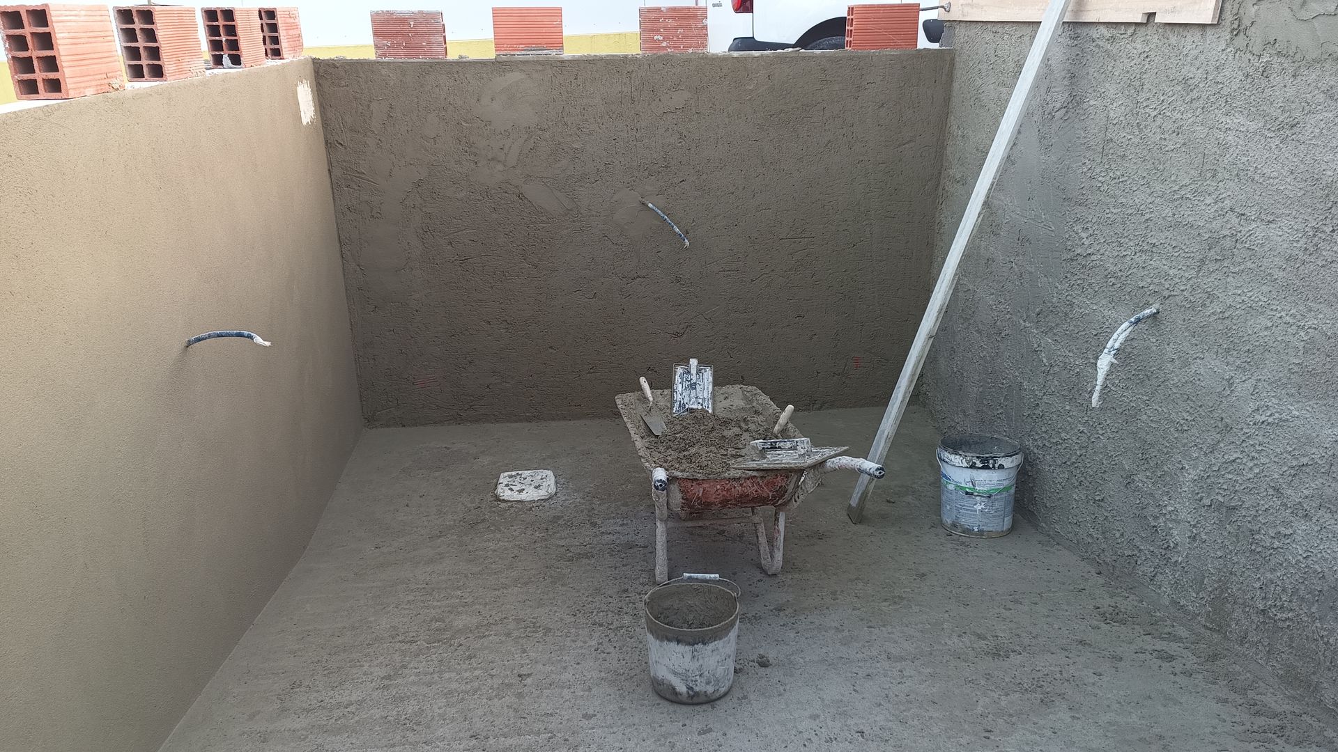 Vista interna de um projeto de construção: um espaço retangular com paredes e piso de cimento, contendo ferramentas e materiais.