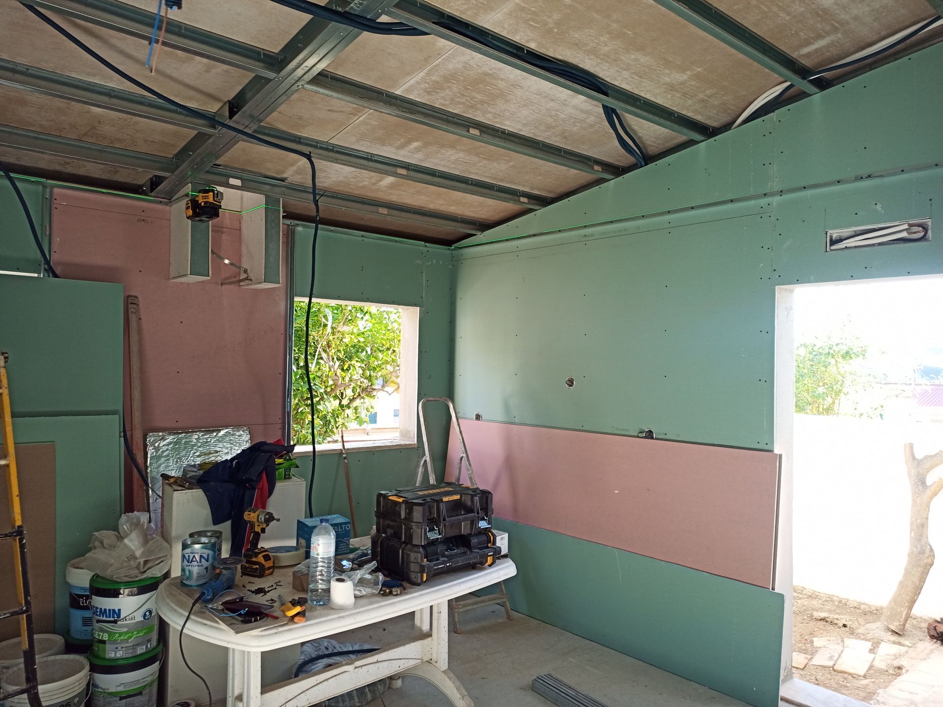 Interior de um cômodo em construção: parede de gesso, estrutura metálica exposta, janela, ferramentas sobre uma mesa e porta aberta.