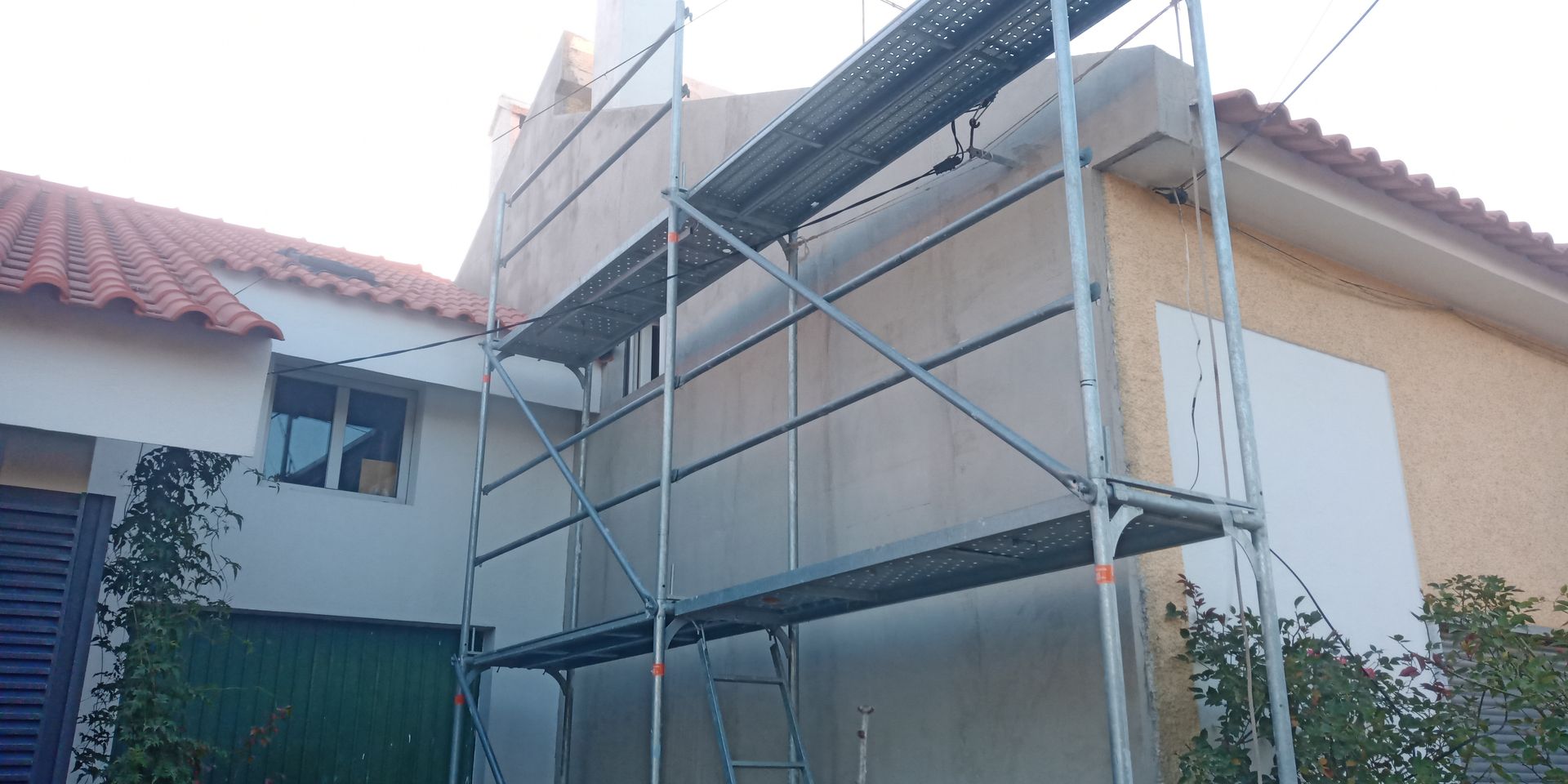 Andaimes montados junto a um edifício com parede amarela e telhado vermelho; possível obra de construção.