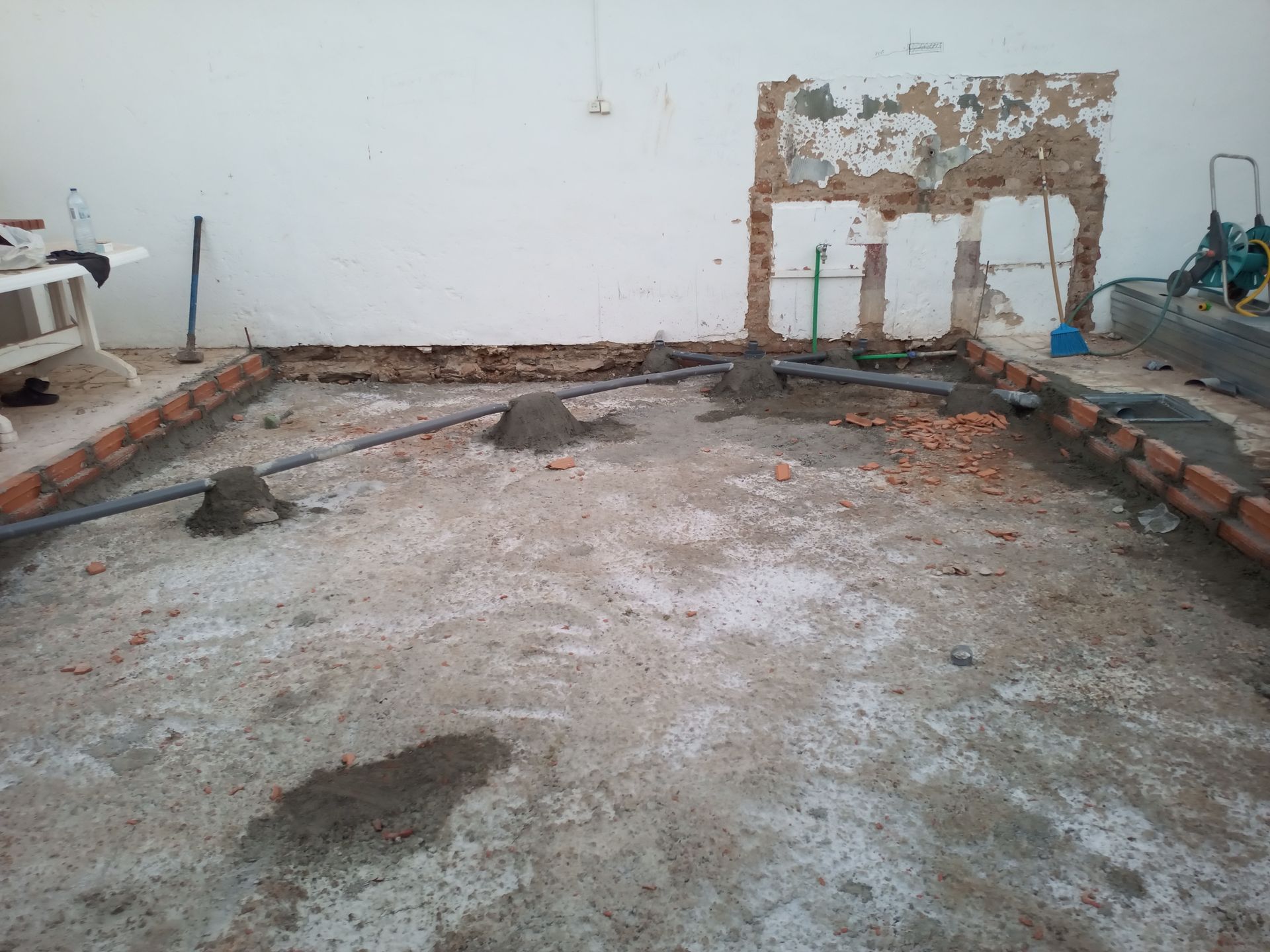 Canteiro de obras: borda de tijolos, piso de concreto, encanamento exposto, parede danificada, materiais de construção.