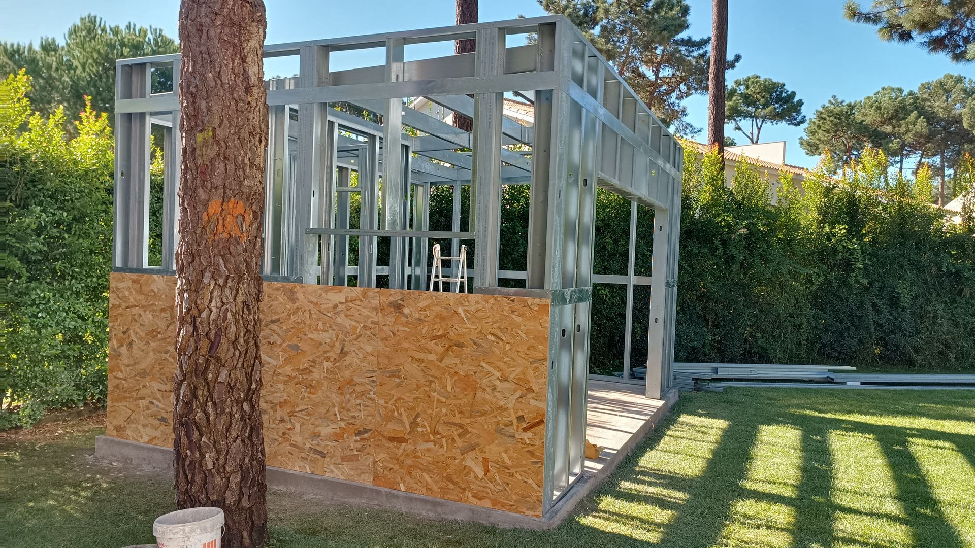 Estrutura metálica de um pequeno edifício em construção, com painéis de madeira na base, assentada na relva, perto de uma árvore.