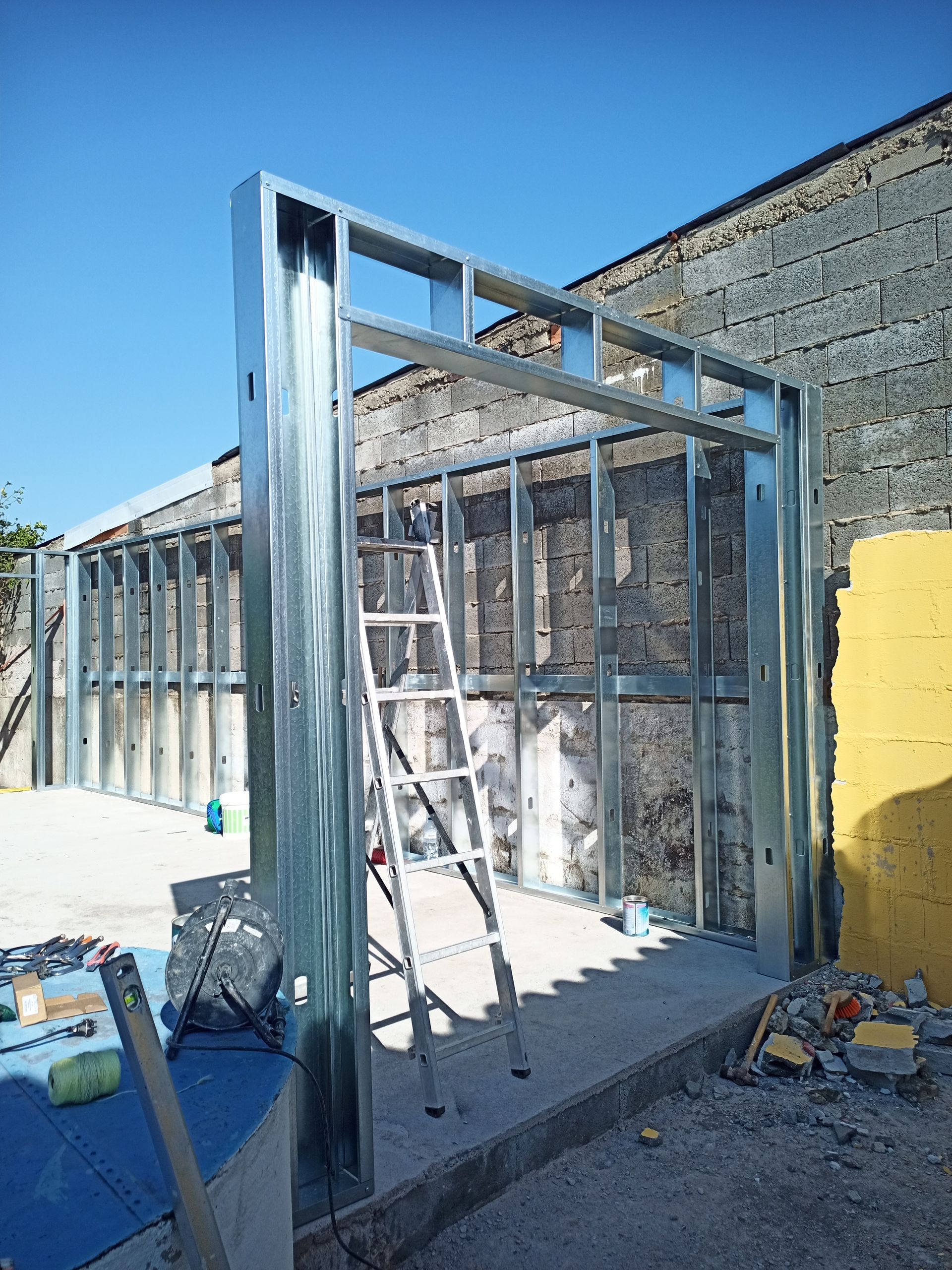 Estrutura metálica da fachada de um edifício, com uma escada encostada nela. Céu azul e parede de tijolos ao fundo.