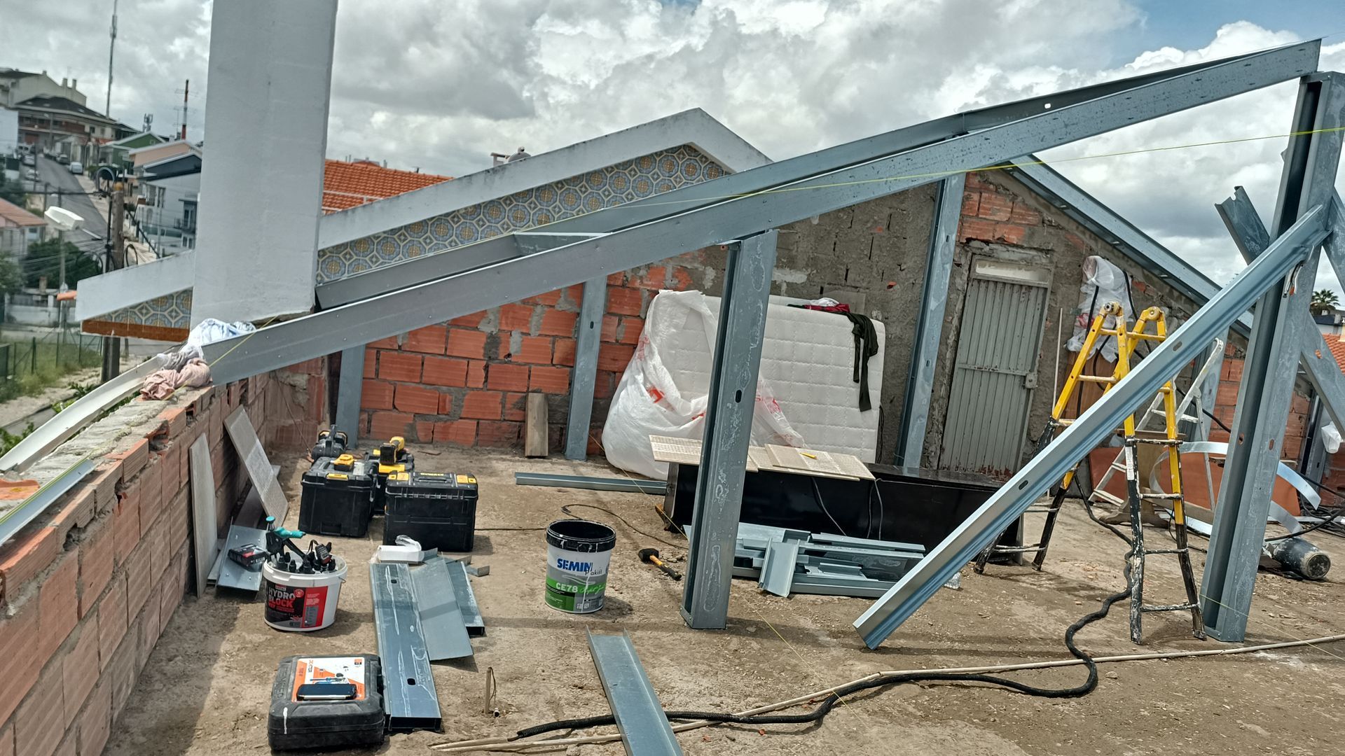 Construção no telhado: Vigas de metal e estrutura de tijolos, ferramentas e materiais espalhados, ambiente iluminado pela luz do dia.