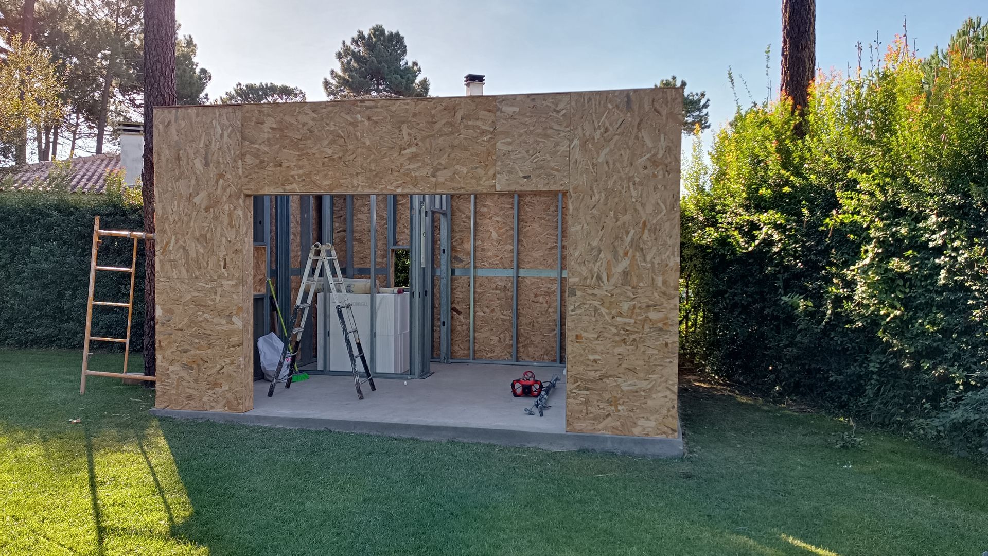 Garagem em construção: paredes revestidas com OSB, porta aberta, ferramentas, escada, base de concreto, gramado verde.