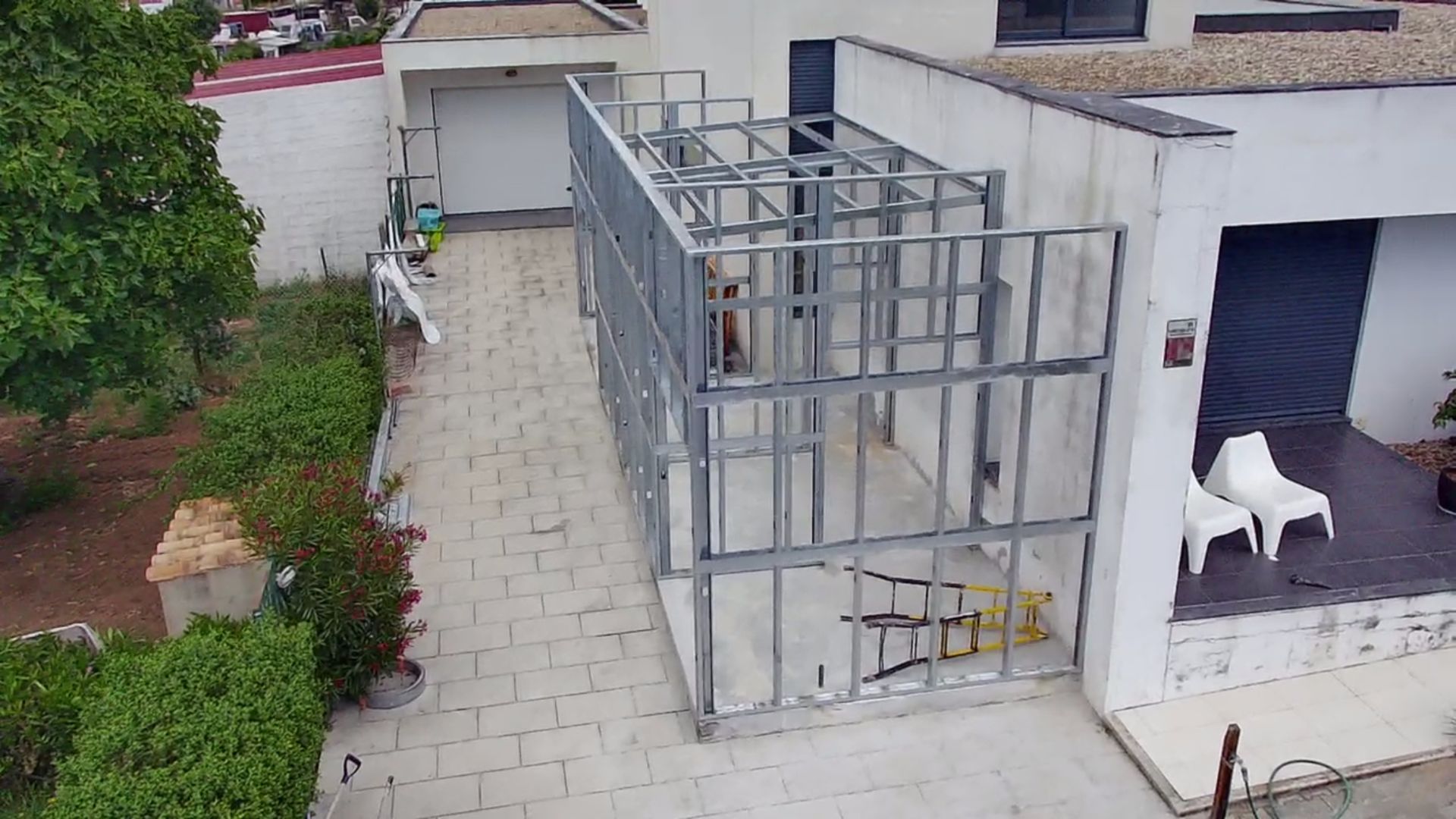 Estrutura metálica de uma extensão de edifício em construção ao lado de uma casa com entrada para carros.