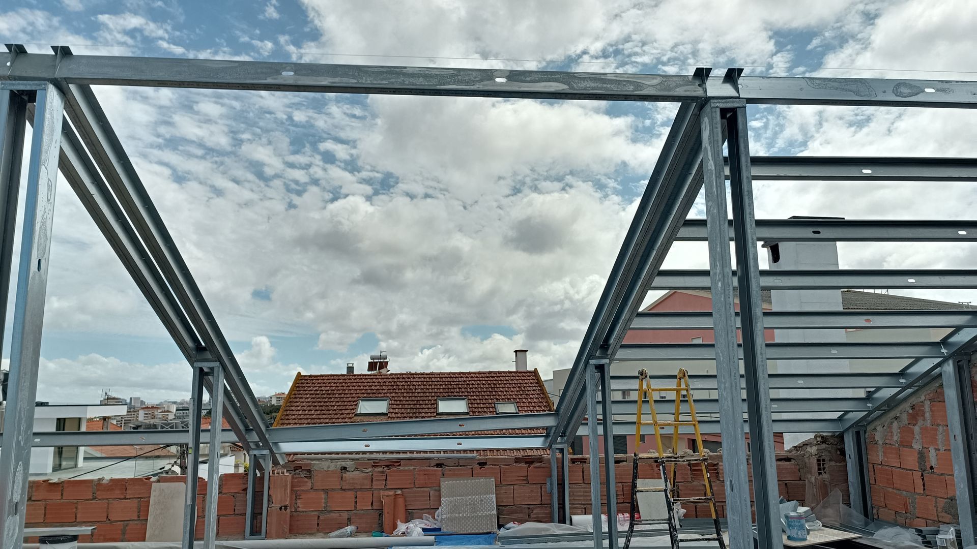 Estrutura metálica de um edifício contra um céu nublado, com telhados visíveis e materiais de construção à mostra.