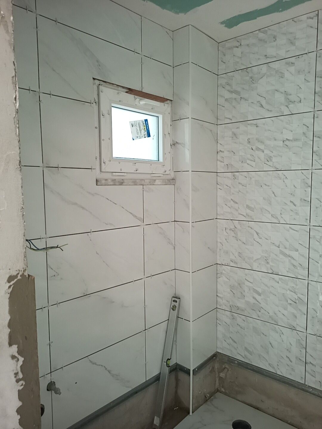 Banheiro em construção com azulejos brancos imitando mármore nas paredes, ao redor de uma pequena janela.