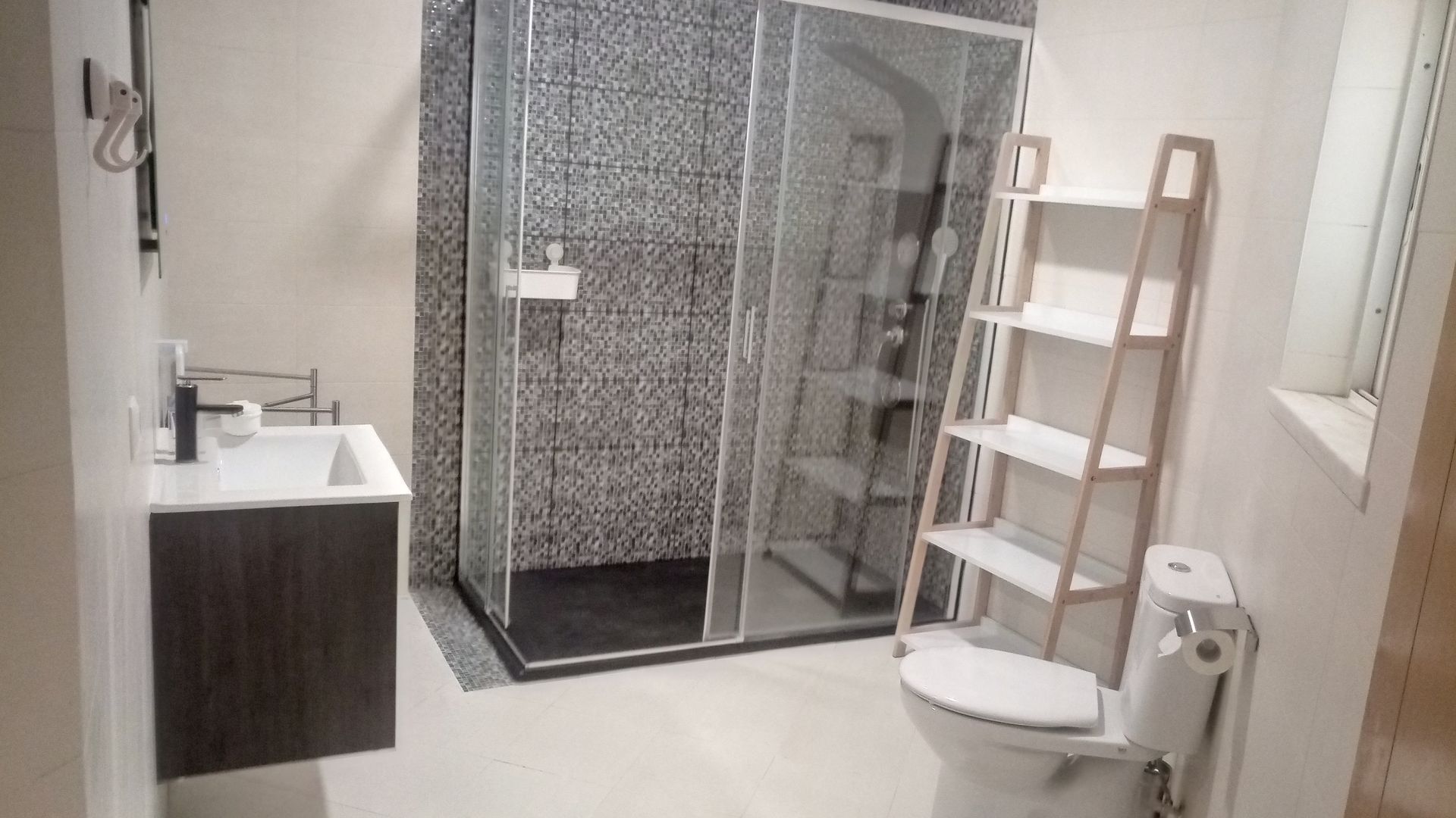 Banheiro moderno com box de chuveiro revestido com azulejos cinza, pia suspensa, vaso sanitário e prateleira branca.