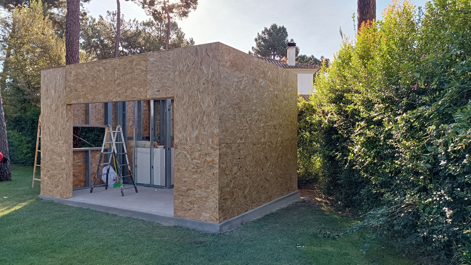 Estrutura inacabada de um galpão feito com painéis OSB sobre uma base de concreto, situado em um gramado com árvores ao fundo.