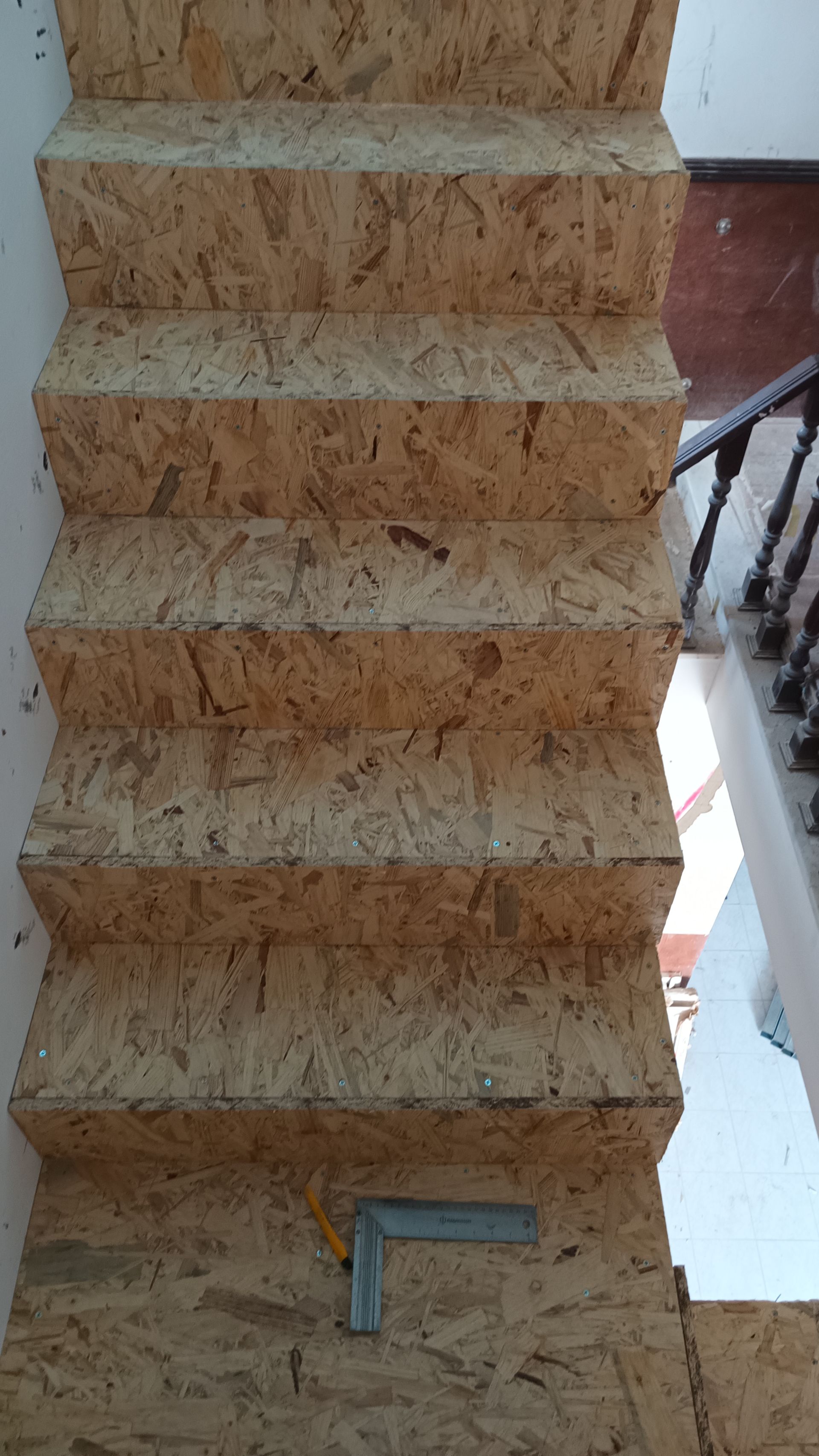 Escada construída em aglomerado de cor clara, com uma ferramenta quadrada de metal apoiada no degrau inferior.